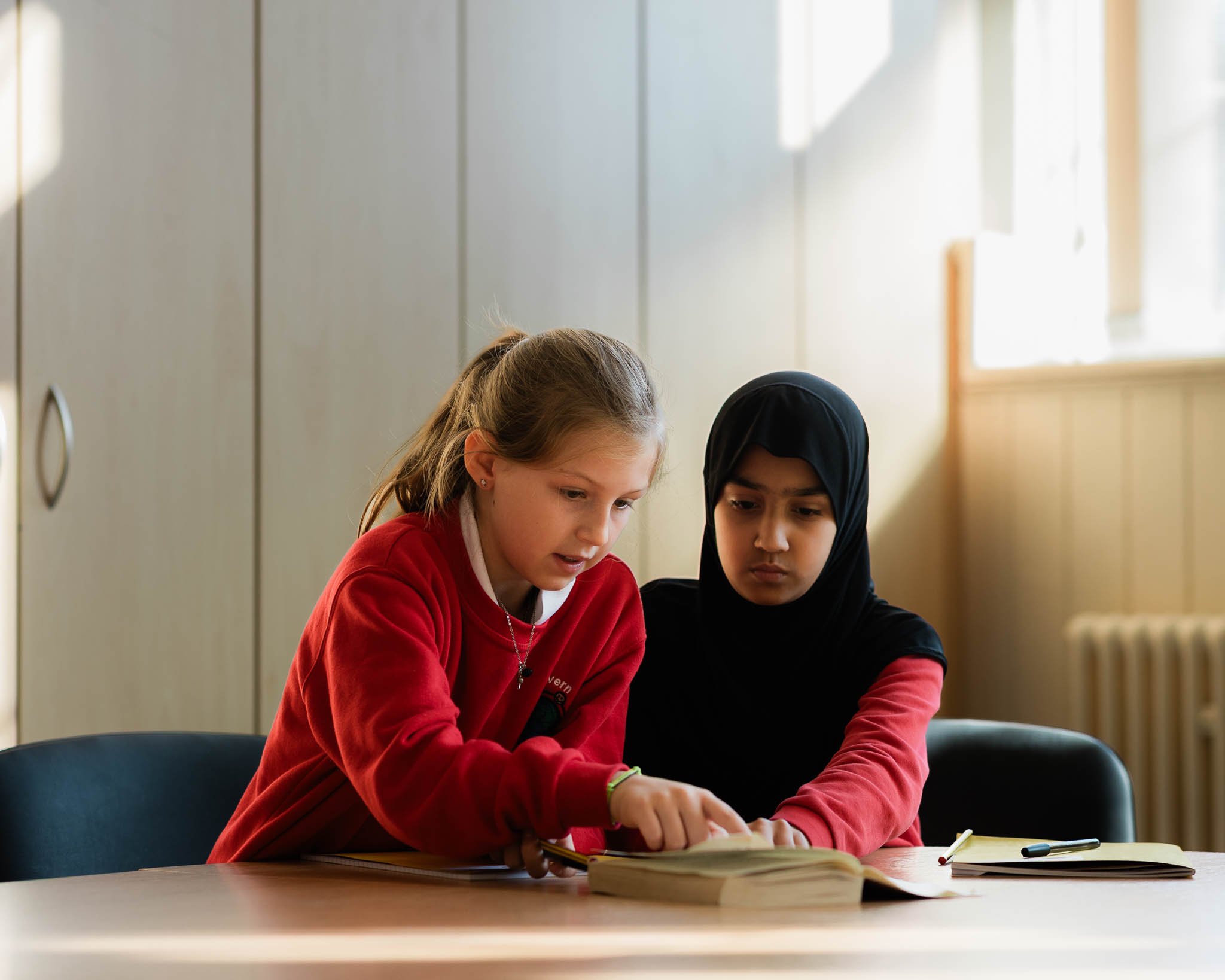 Two young girls, one with light skin and the other with darker skin wearing a hijab, sitting at a table and reading a book together in a room with beige walls and a window.