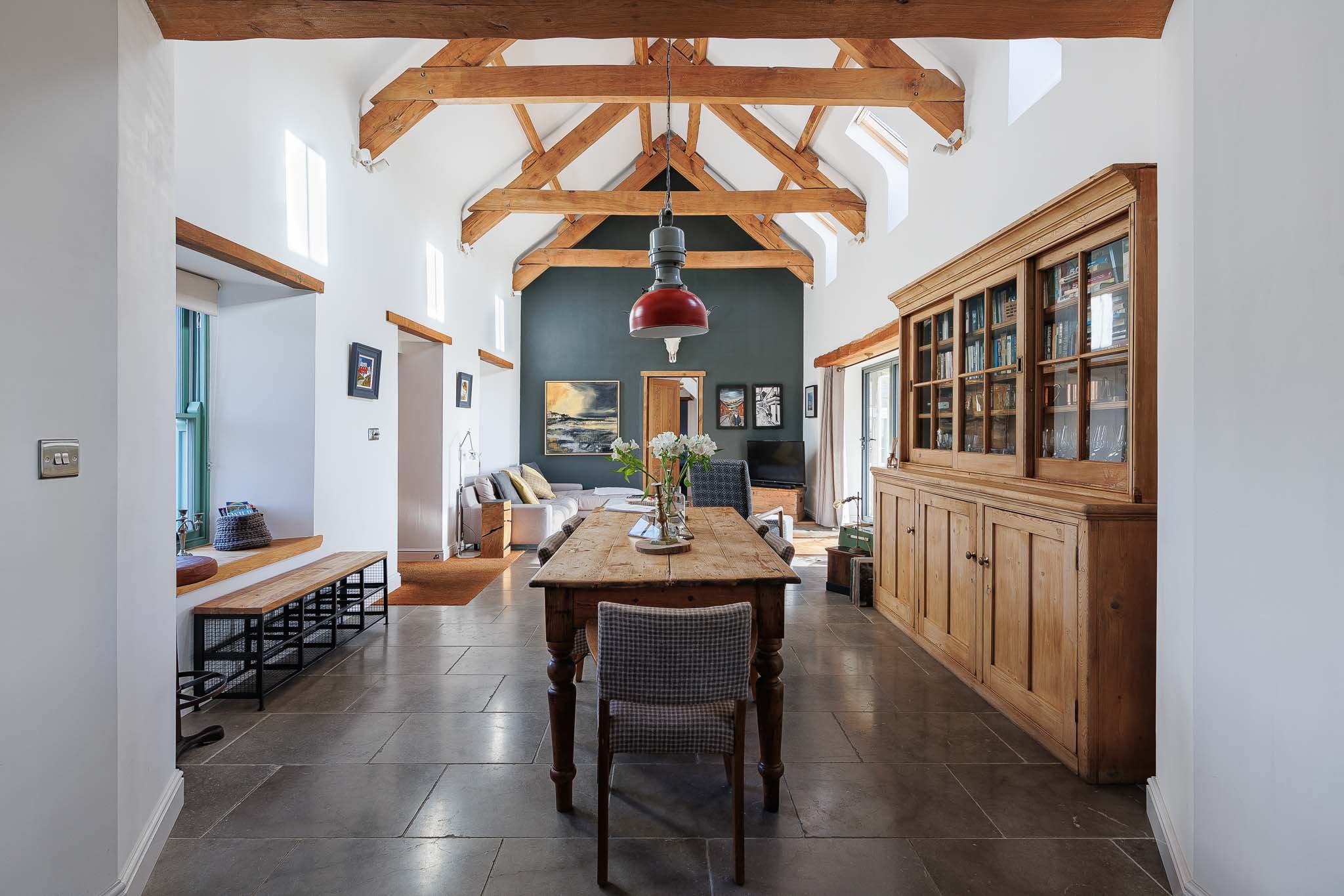 Living room with vaulted ceiling featuring wooden beams, a dining table with chairs, a wooden bookcase, a gray accent wall with art, and windows allowing natural light.