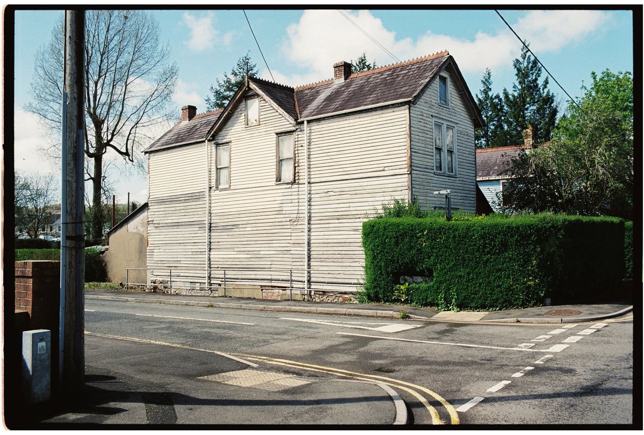 An old, white wooden house with a gabled roof and multiple windows next to a green hedge and a street with painted lines and a curb, under a partly cloudy sky.