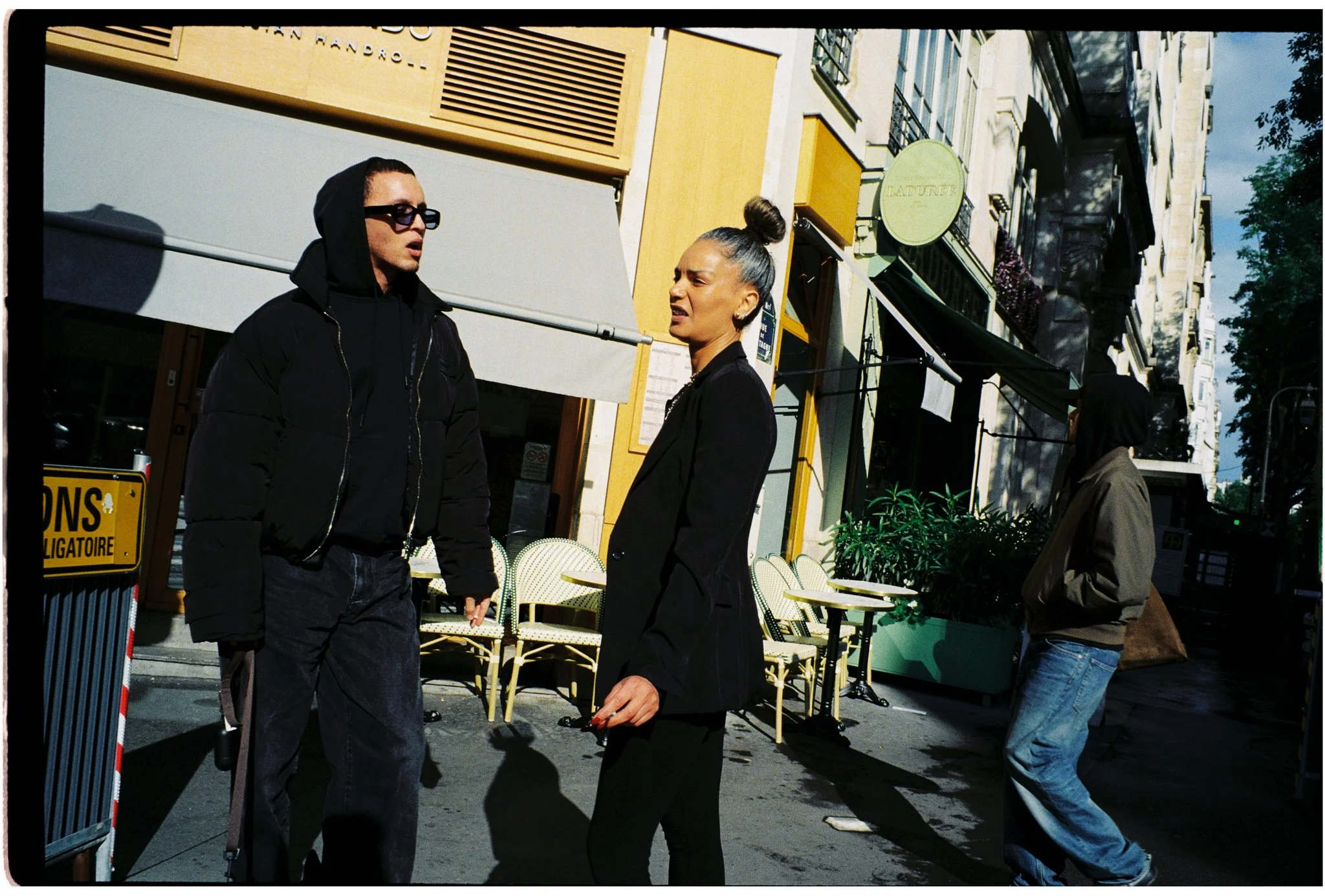 Two people, a man wearing sunglasses and a woman with a top bun, are standing outdoors on a city street, engaged in conversation. Behind them are outdoor cafe chairs, a green sign, shops, and multi-story buildings with balconies. The man is dressed i