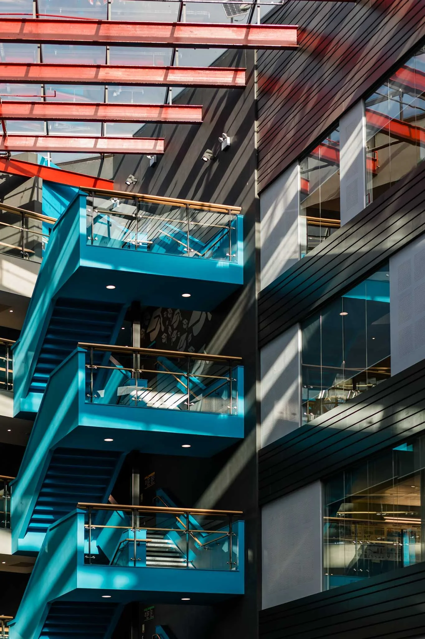 Multistory modern building interior with black and wood-paneled walls, blue and red staircases with glass railings, sunlight casting shadows through windows.