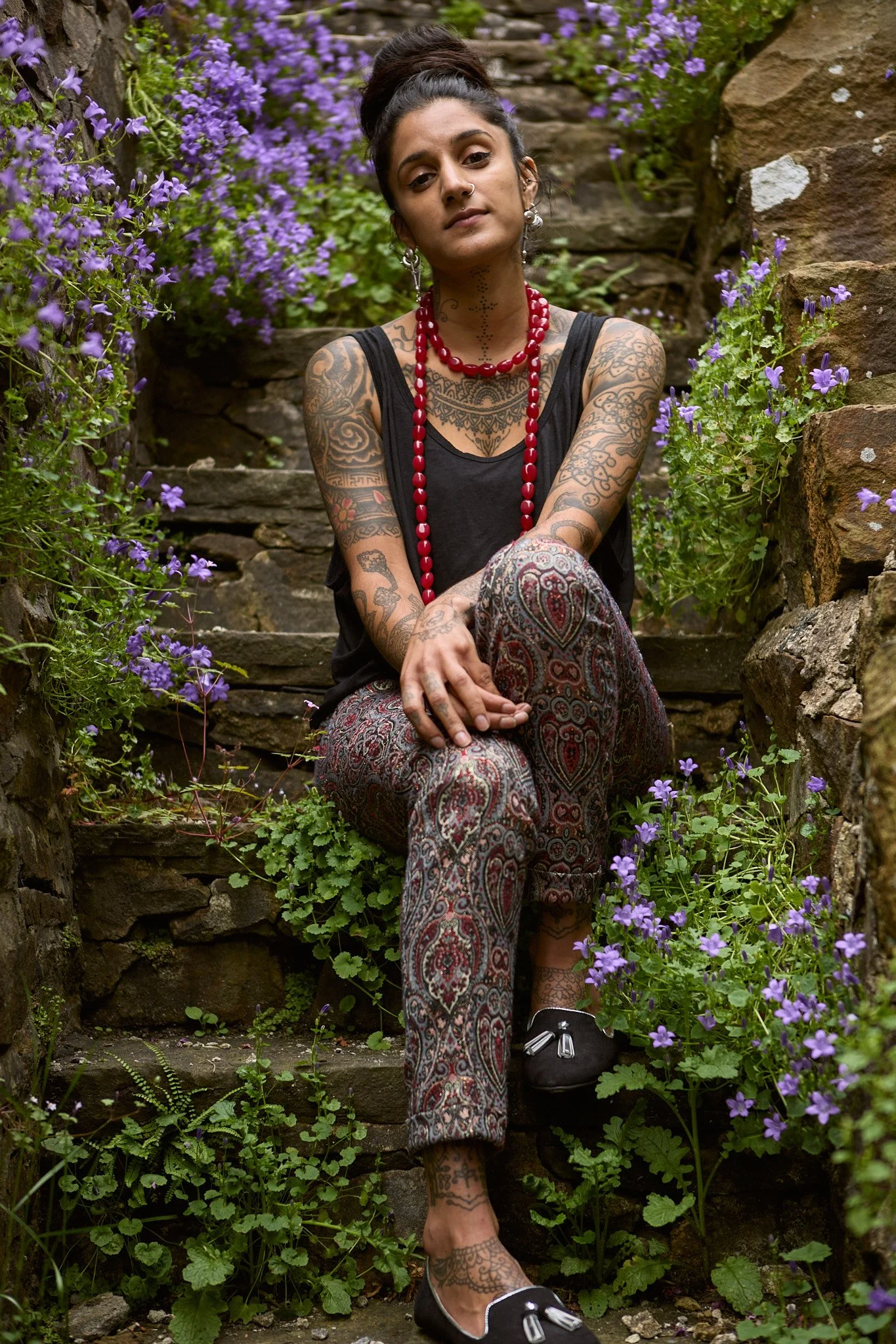 A woman with tattoos and dark hair in a bun sitting on stone steps surrounded by purple flowers, wearing a black tank top, patterned pants, and black slip-on shoes with bows.