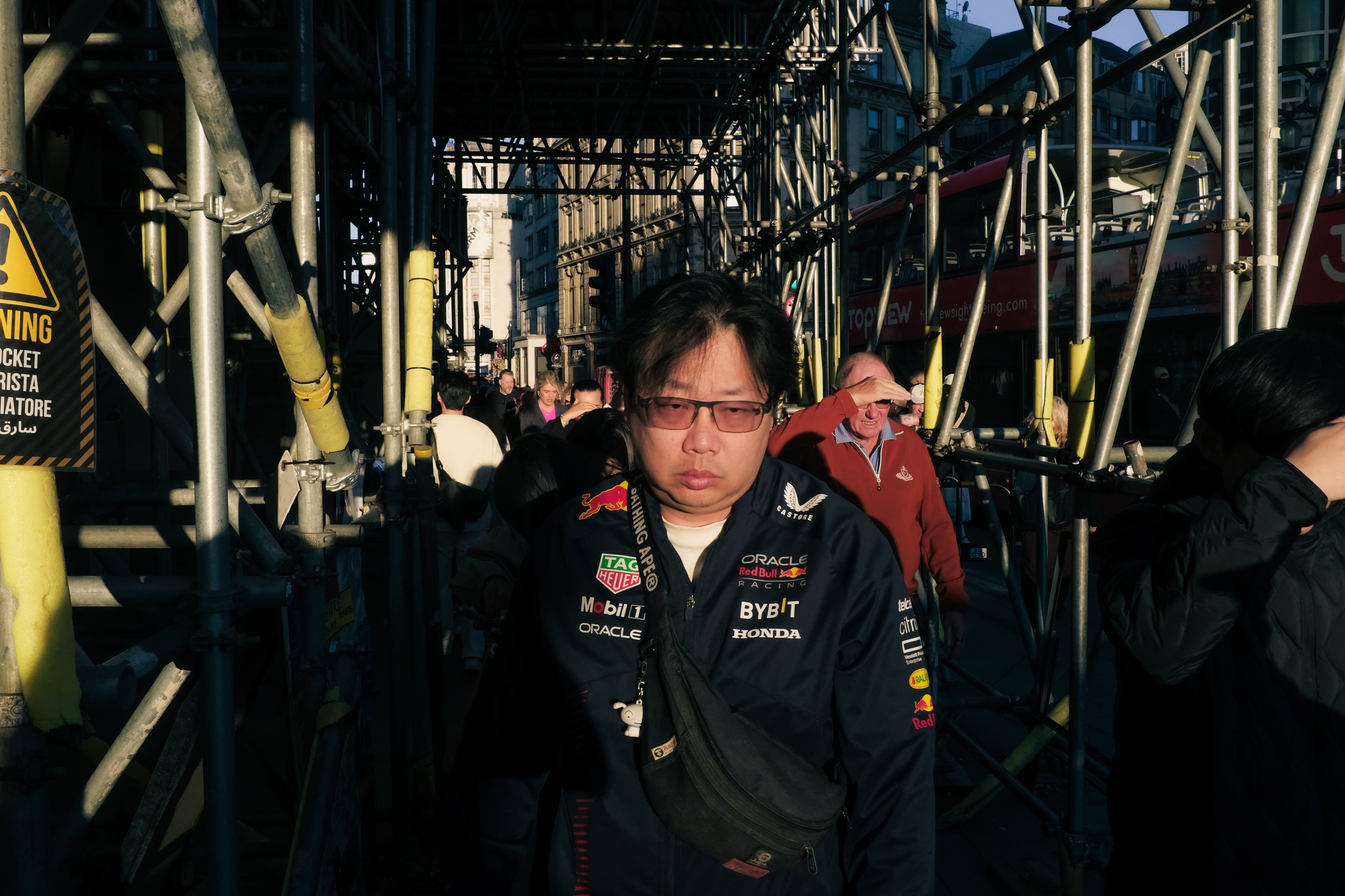 A man wearing a racing jacket with sponsor logos, including Red Bull and Honda, stands among construction scaffolding on a city street. Several people are in the background, and a double-decker bus can be seen.