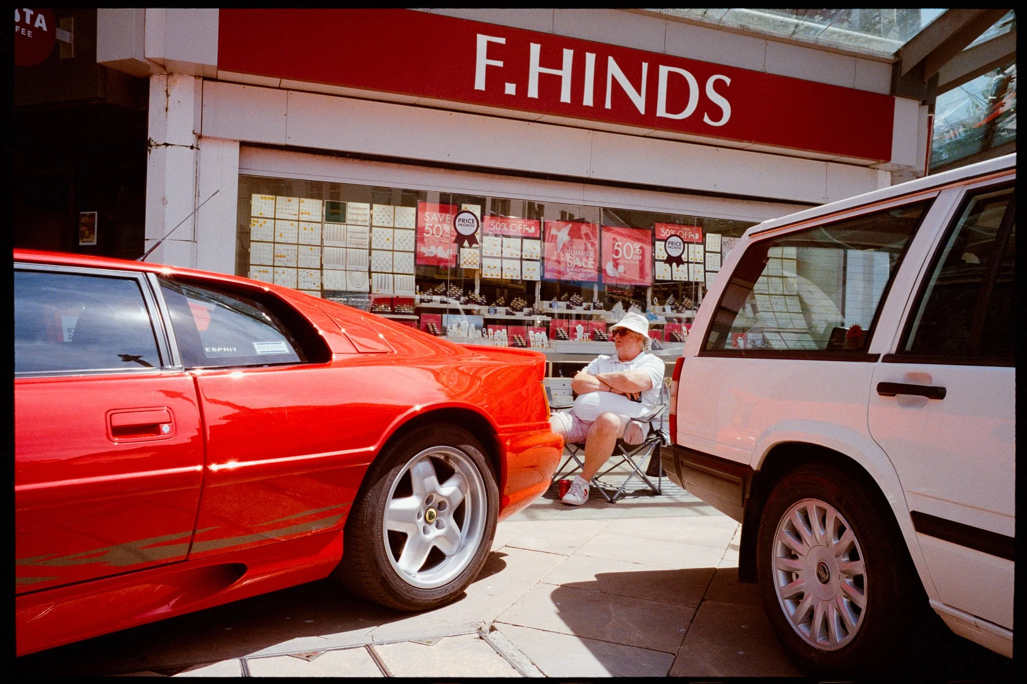 A man sitting in a foldable chair between two parked cars in front of a store with a large red sign that says 'F. HINDS.' The man is wearing a white bucket hat, sunglasses, a white shirt, and red sneakers. The store window displays signs indicating s