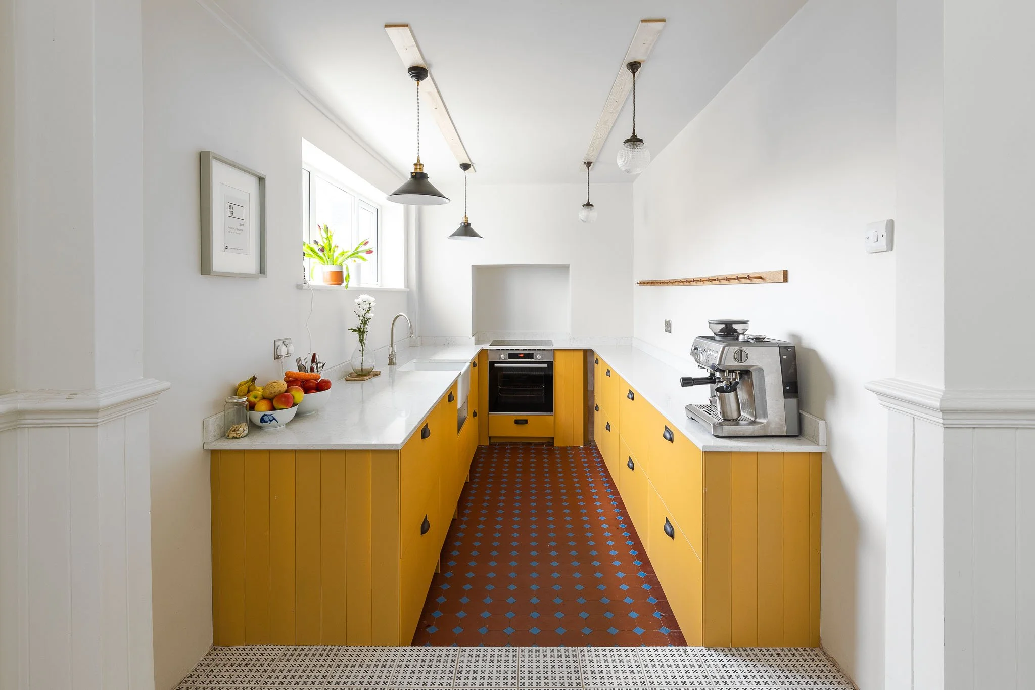White kitchen with yellow cabinets, black hardware, and a red and blue patterned tile floor. Features include a window with a plant, fruit bowl, and coffee machine.