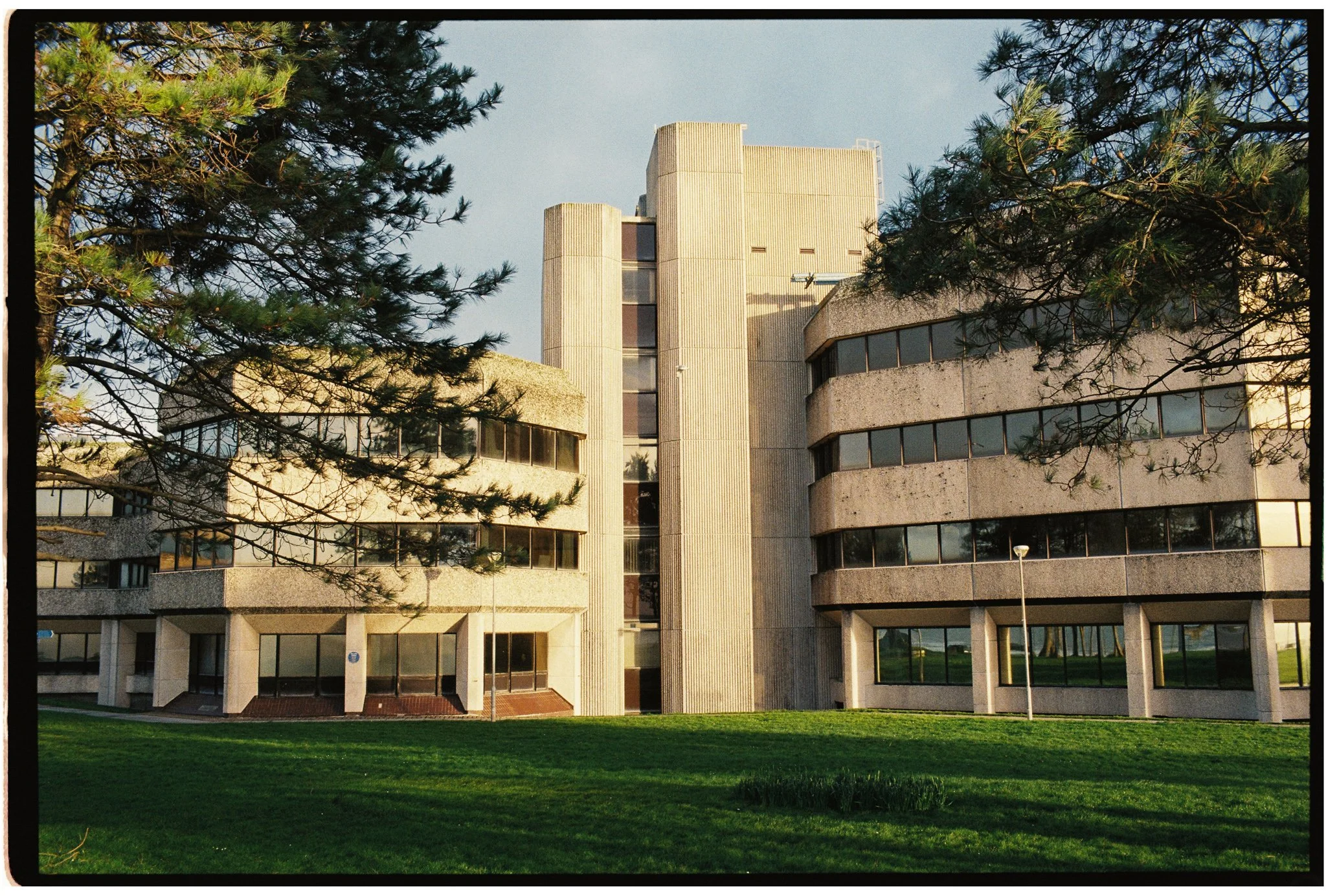 Modern concrete building with curved floors, surrounded by green grass and trees, under a pale blue sky.