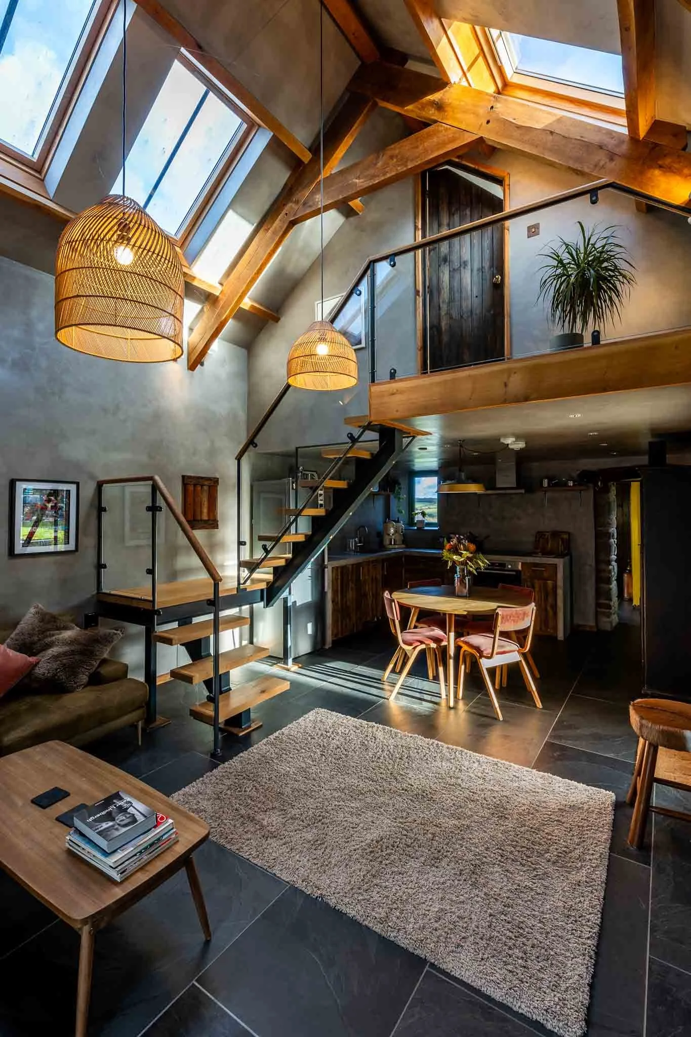 Interior of a modern loft with high ceilings, wooden beams, large skylights, a small round dining table with pink chairs, a cozy sofa, a staircase leading to an upper level, and contemporary decor.