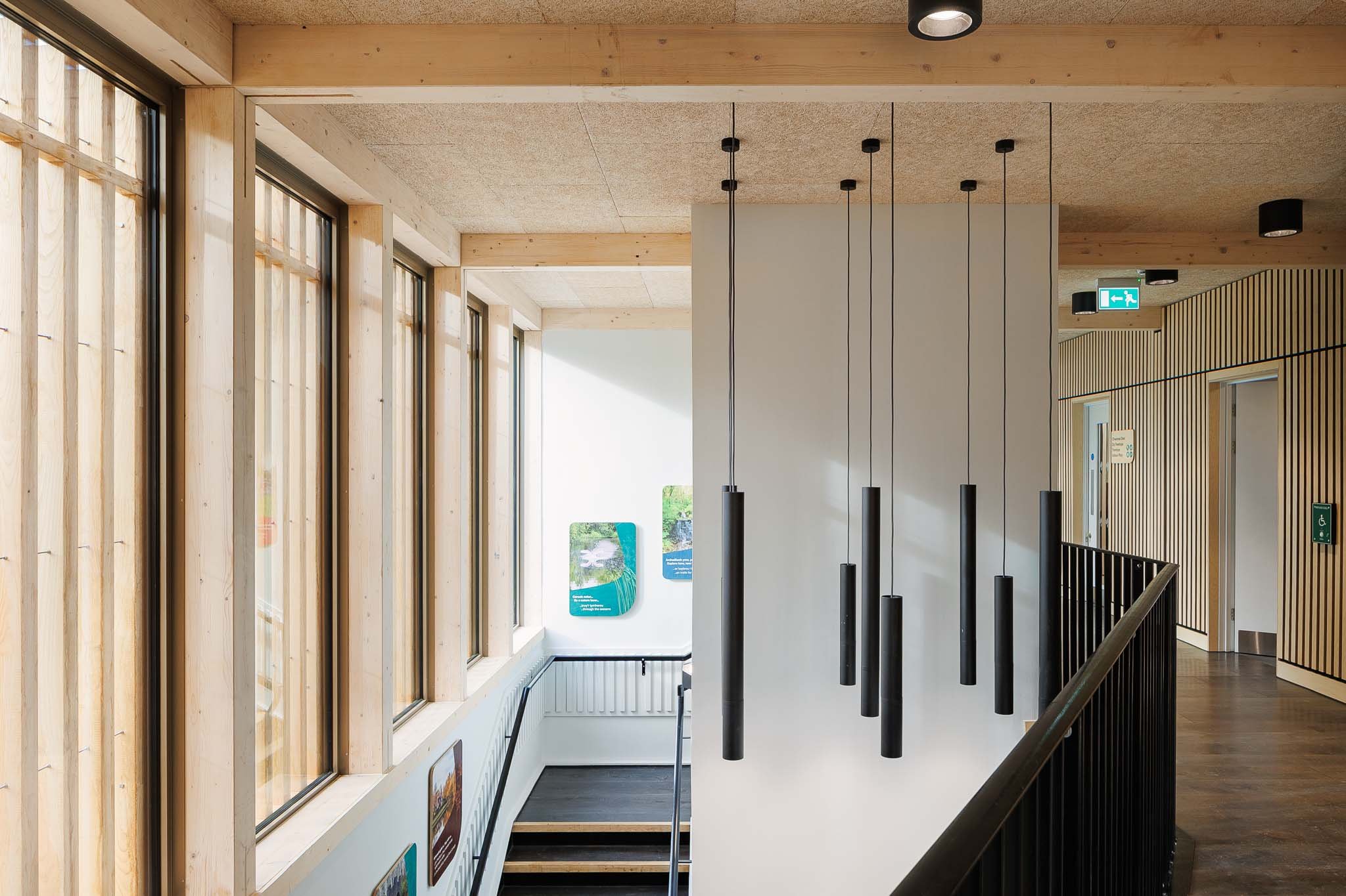 Interior view of a modern building with large windows, wooden wall paneling, ceiling with black circular recessed lights, and black hanging pendant lights. There is a staircase with a black handrail, and signs indicating an emergency exit and accessi