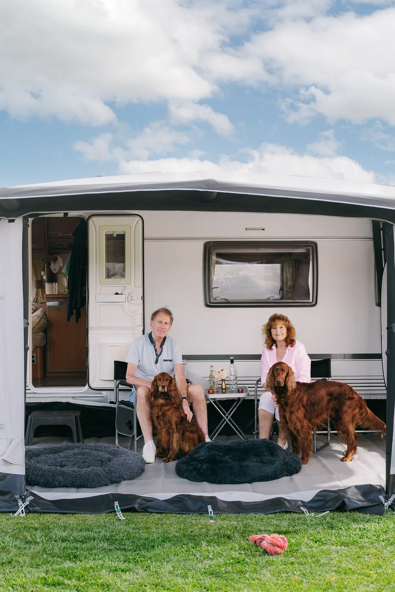 A couple sitting outside a mobile home with their two golden retrievers. The man is wearing a light blue shirt and shorts, the woman is wearing a pink blouse. There are two fluffy black rugs on the ground, a small table with drinks and flowers, and a