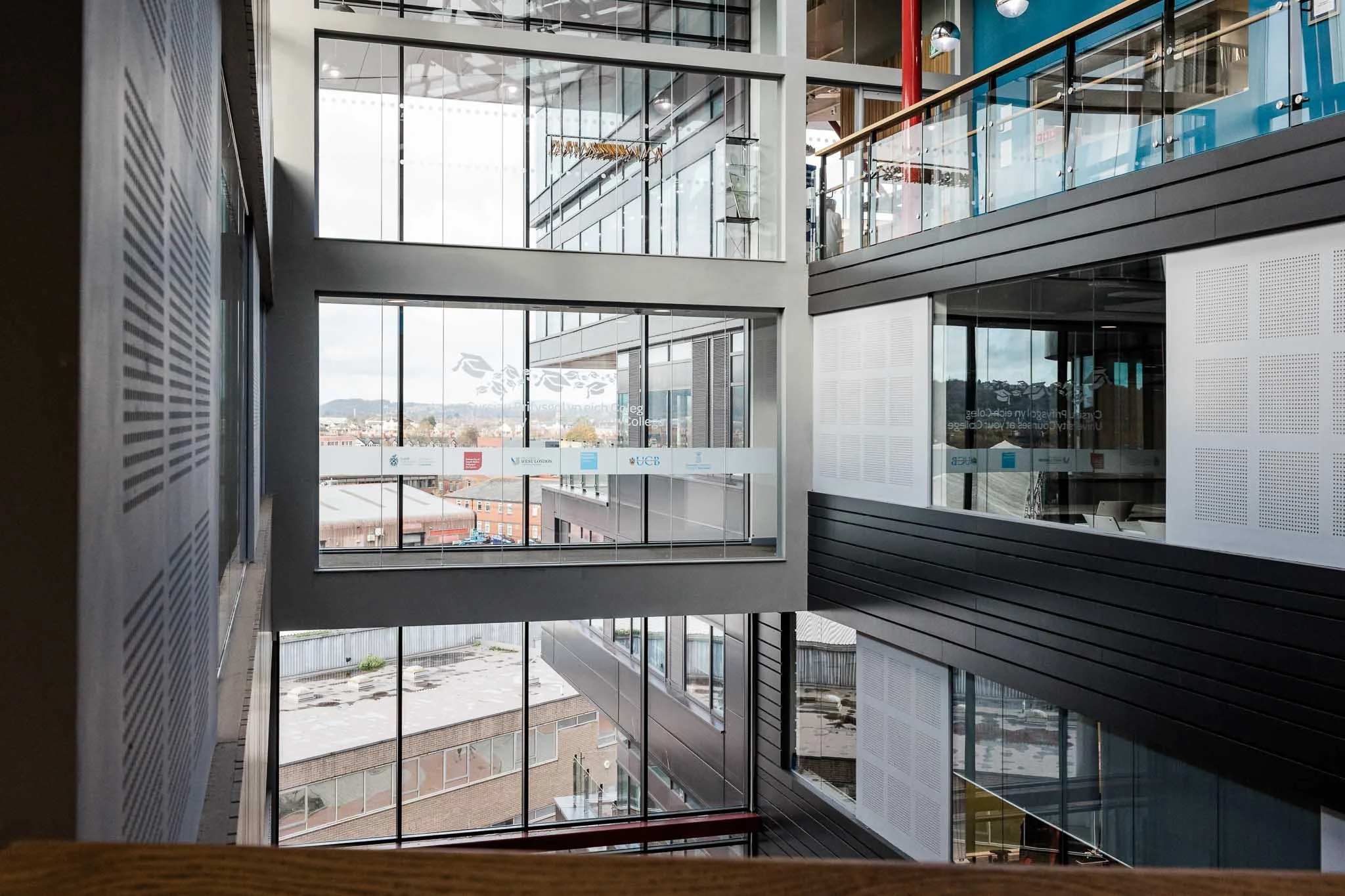 Interior view of a modern multi-story building with large glass windows showing a cityscape and hills outside. The photo is taken from a higher floor looking down at the lower levels, featuring glass railings and a minimalist design.