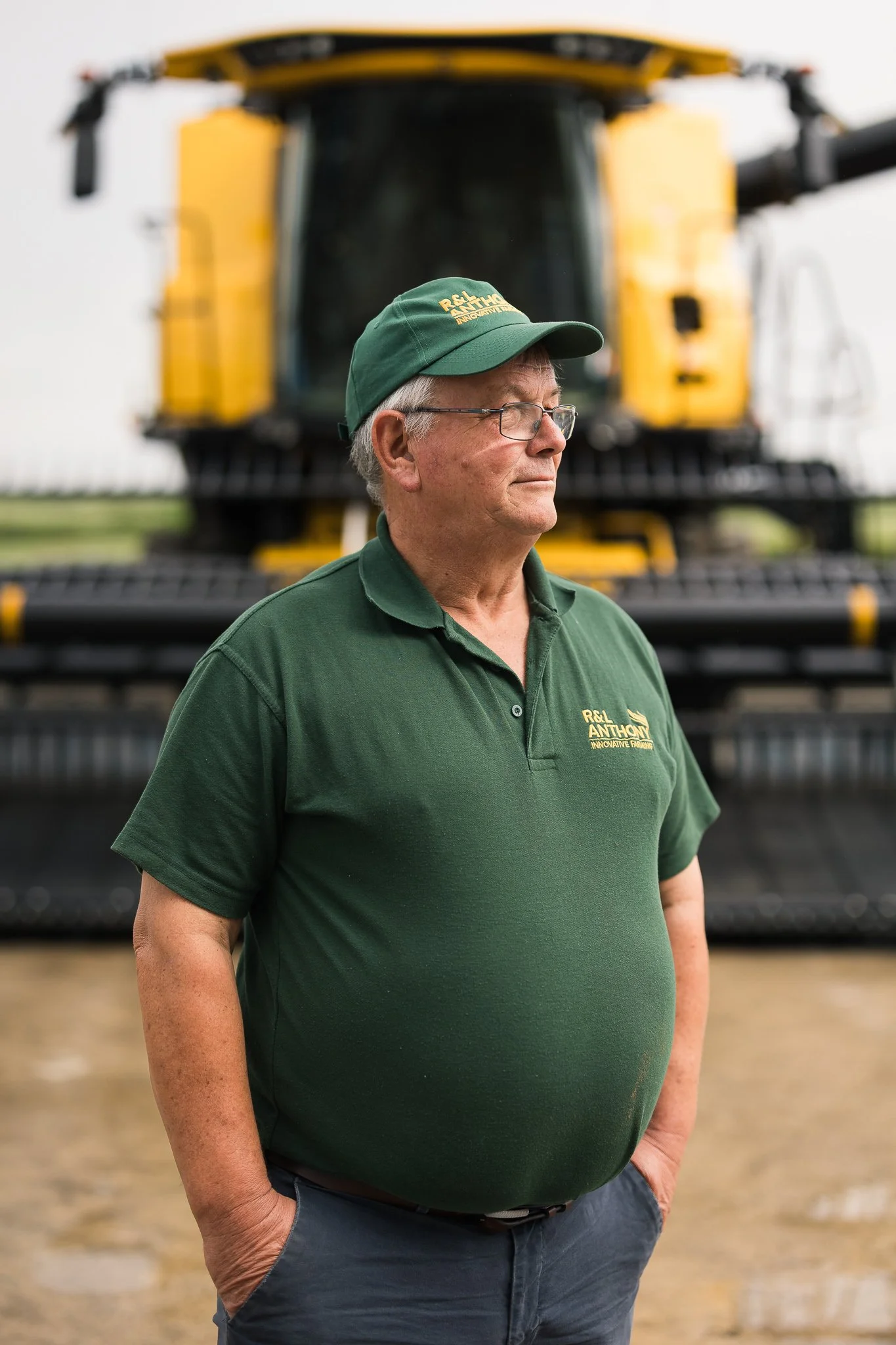 A mature man wearing glasses, a green cap, and a green polo shirt with logo details, standing outdoors in front of a large yellow machinery, with hands in pockets and looking to the side.