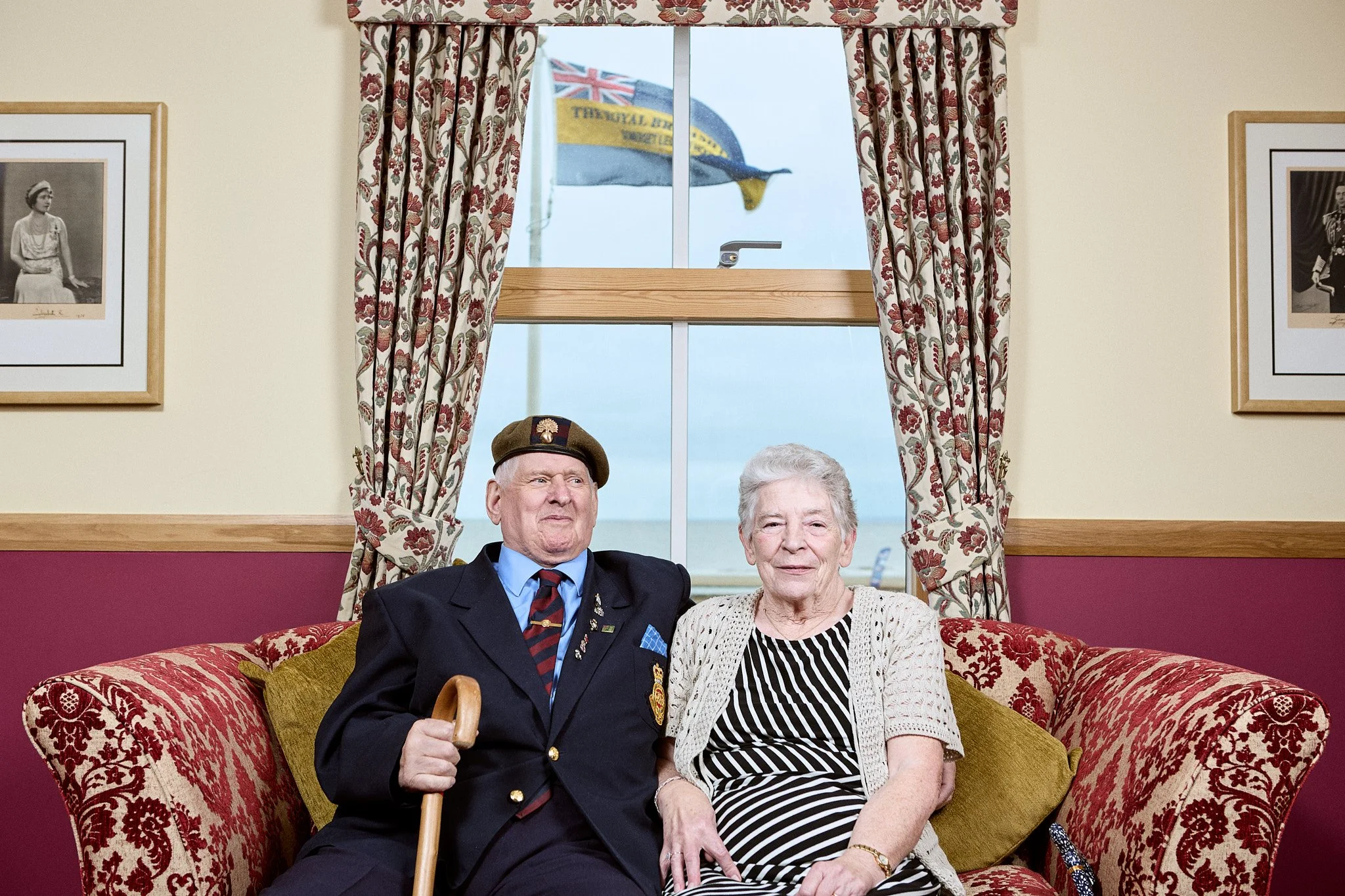 An elderly man in a military uniform and an elderly woman sitting on a red floral-patterned sofa in a living room with cream-colored walls, red wainscoting, and floral curtains. Outside the window, two flags are visible, including a Union Jack flag.