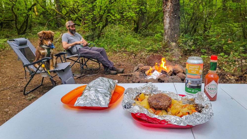 Closeup of a classic foil packet meal with a man and a dog sitting around a campfire in the background