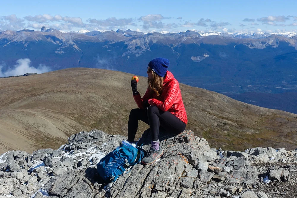 A hiker with a day pack sitting on a rock to eat an apple with mountains in the background.
