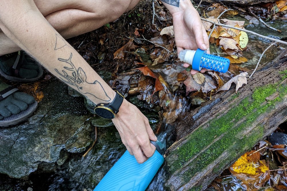 A hiker filling the Hydrapak Flux bottle from a stream - the Katadyn BeFree water filter is in their other hand