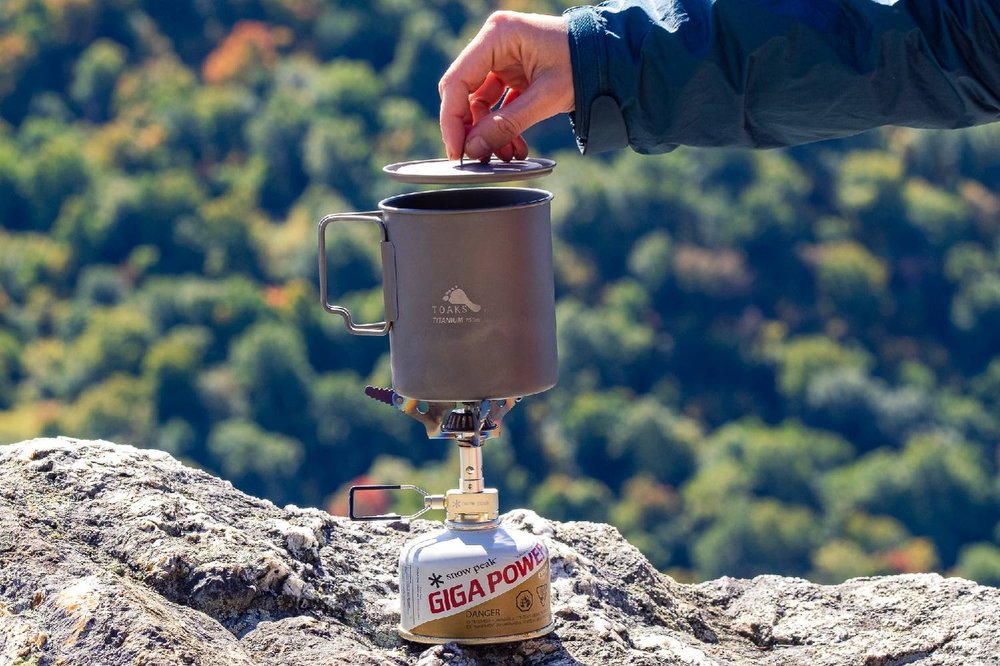 A hand lifting the lid of the Toaks 750 mL pot sitting on top of the Snow Peak Litemax stove