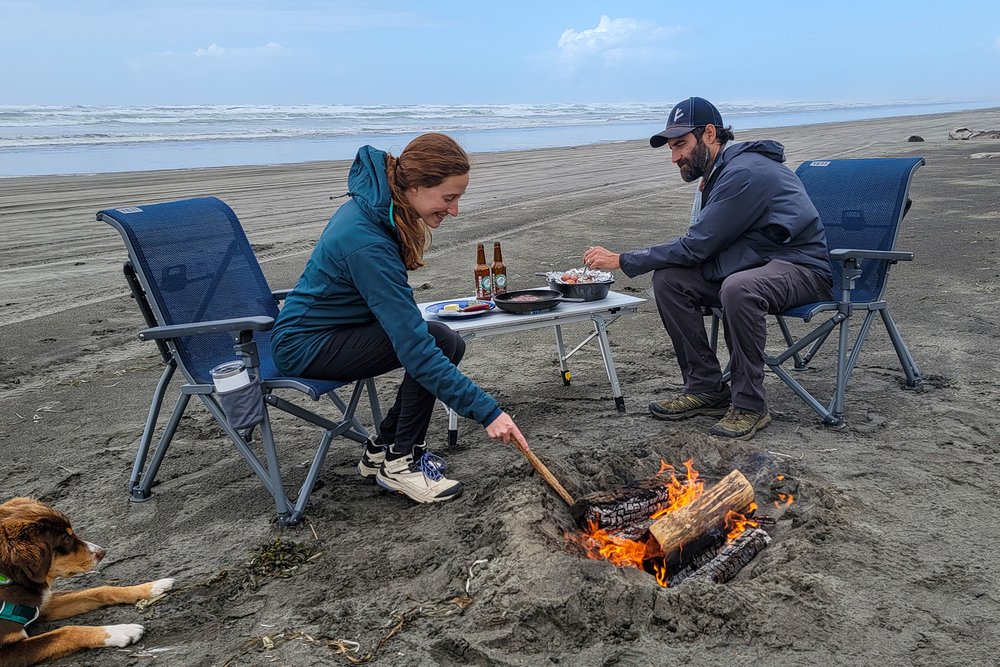 Two people sitting in camp chairs next to a campfire on a beach