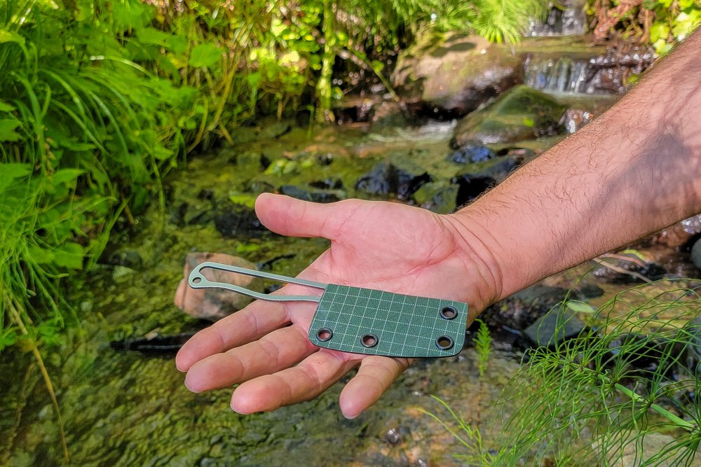 Closeup of the ULA Alpha Knife in the palm of a hikers hand in front of a creek