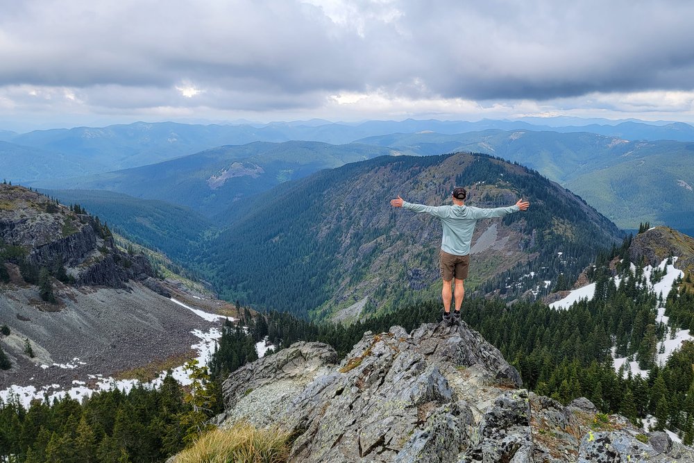 A hiker looking down from the summit of a mountain with their arms spread like a bird
