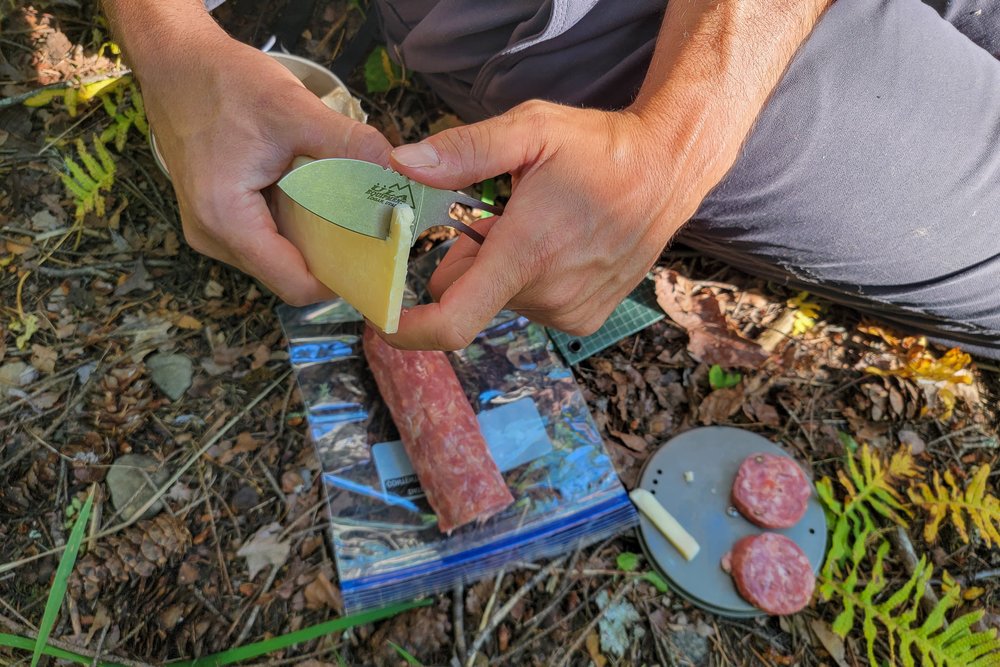 Closeup of a backpacker using the ULA Alpha Knife to cut salami and cheese