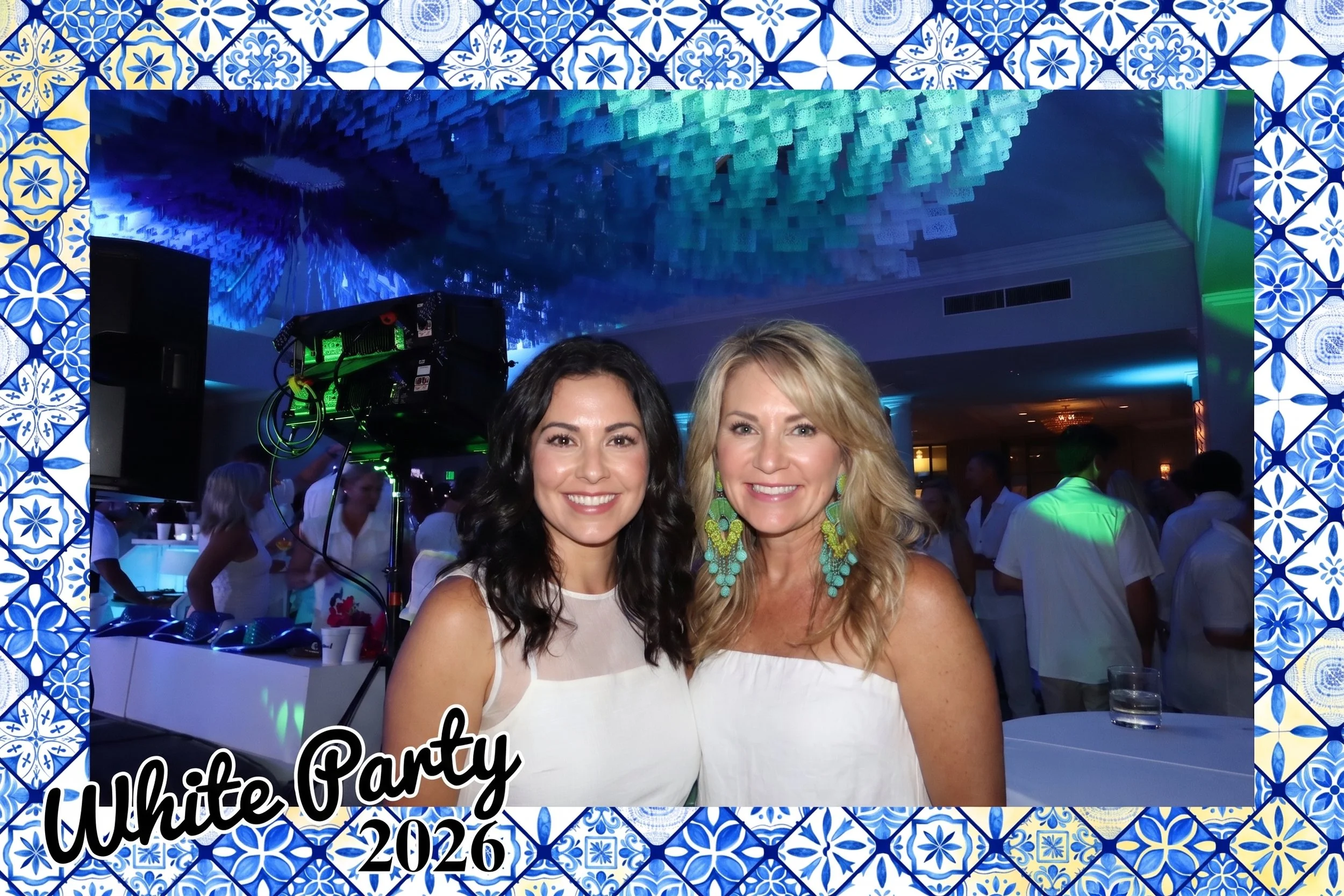 Two guests in white pose in a dark party room lit only by the roaming camera's on-camera flash, framed by a custom blue and yellow tile overlay.