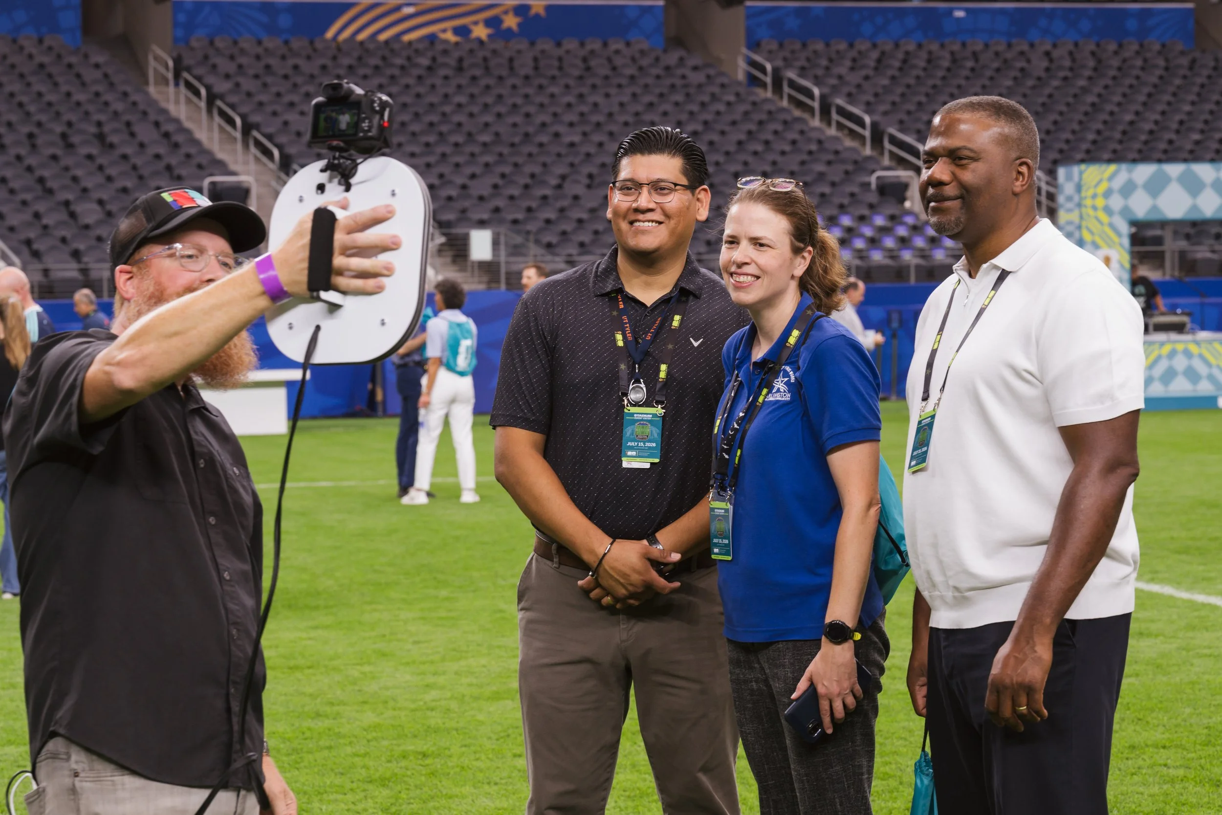 A photographer holds the handheld camera out to photograph three guests standing on a stadium field, their pose showing live on the screen, at a roaming photo booth rental in Dallas-Fort Worth.