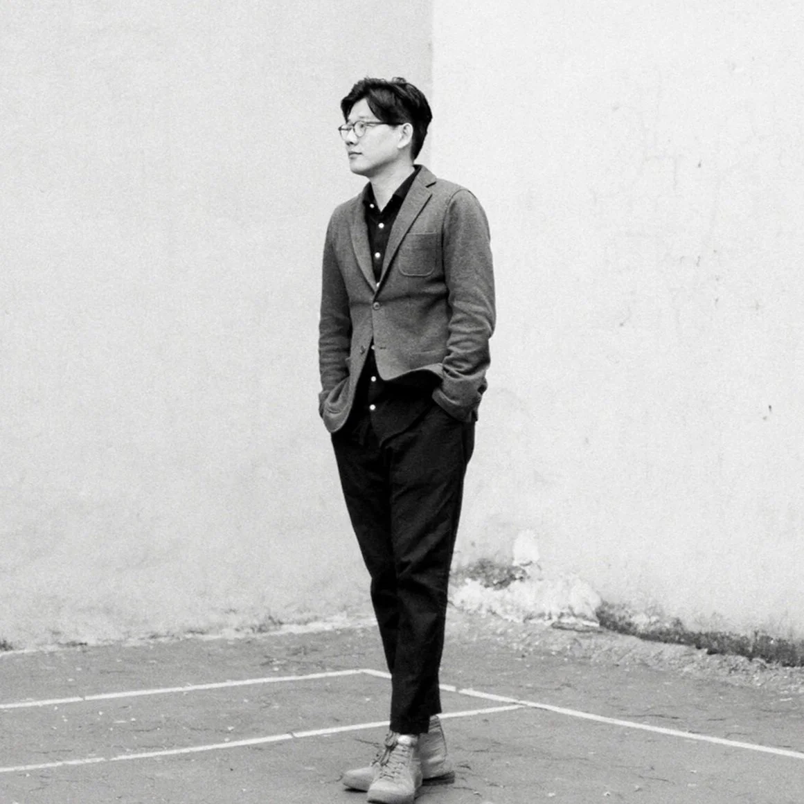 A man standing against a plain wall, dressed in business casual attire with glasses, in black and white photograph.