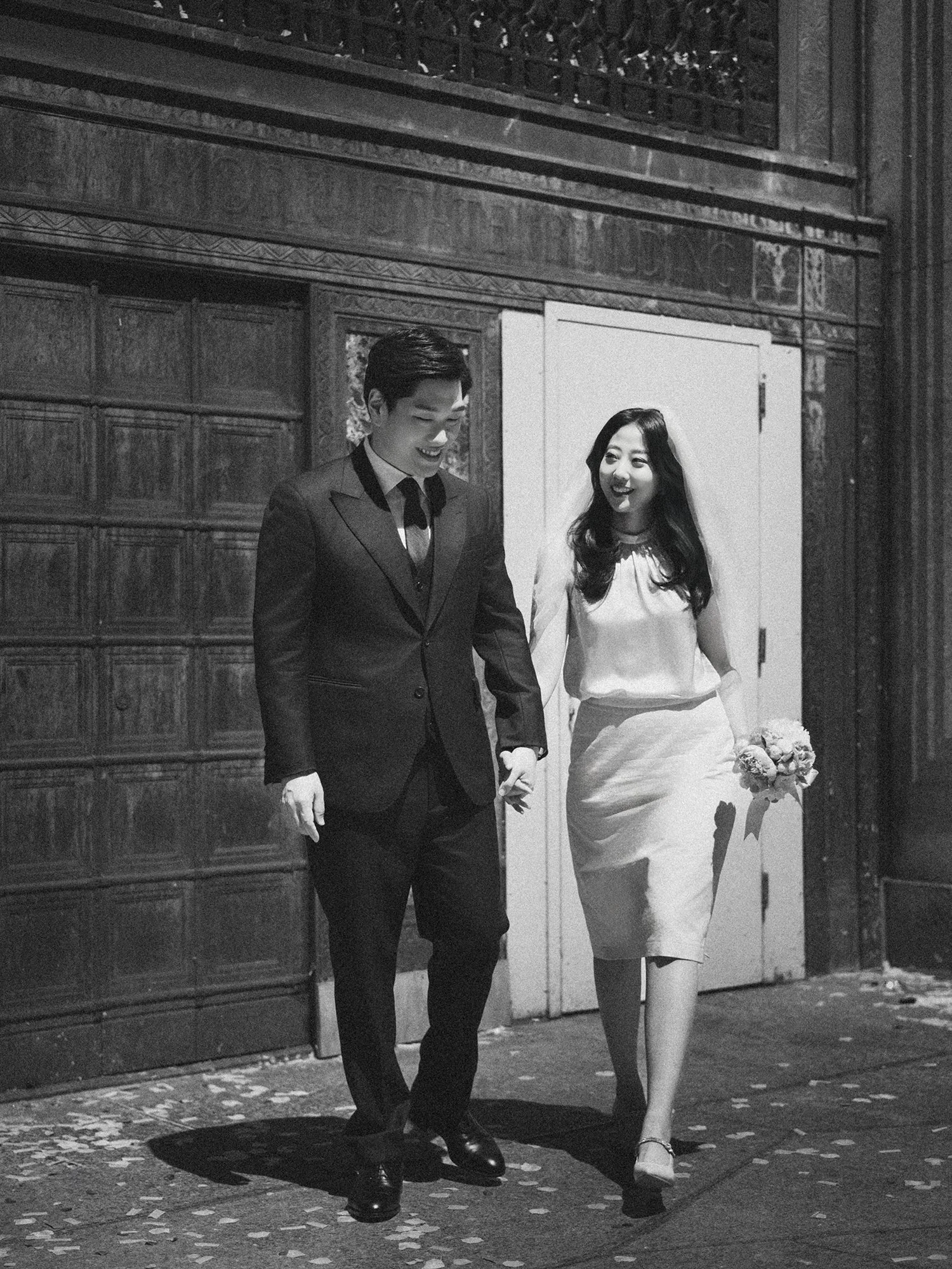 Black and white editorial portrait of a bride and groom holding hands and looking at each other while walking through a historic stone archway in New York City.