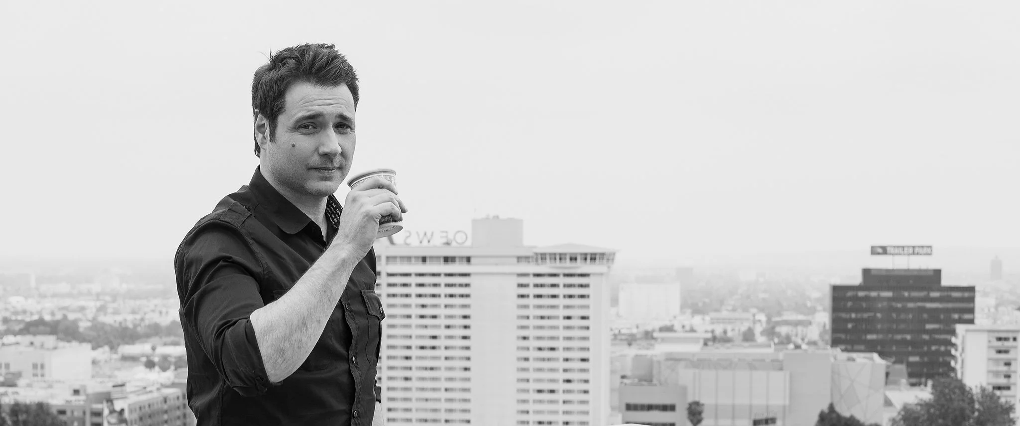 A man in a black shirt holding a cup on a rooftop with a cityscape in the background.