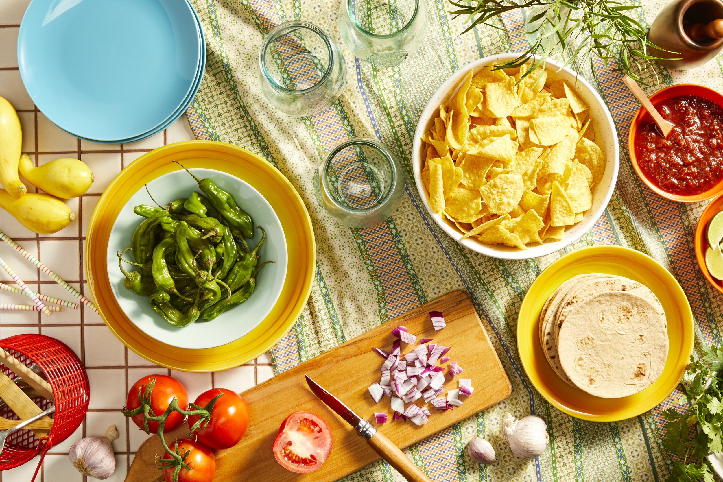 A variety of Mexican food ingredients including bowl of chips, tortillas, shishito peppers, fresh tomatoes, chopped red onion, garlic, salsa, limes, and herbs.