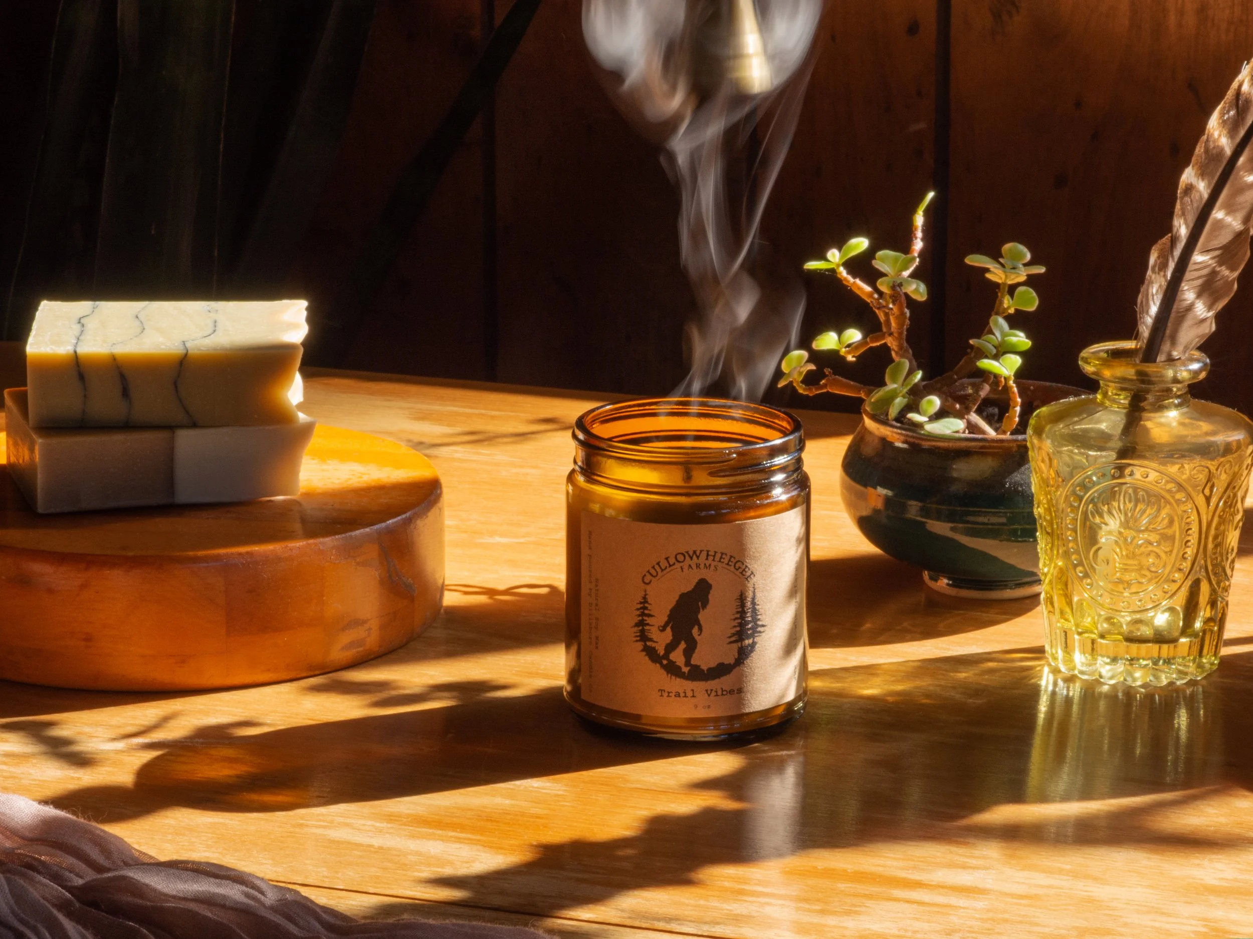 Arrangement on a wooden table including a stack of artisanal soap, a steaming candle in an amber jar labeled 'Cullowhee Farms Trail Vibes,' a small potted plant, and a decorative glass bottle with a feather.