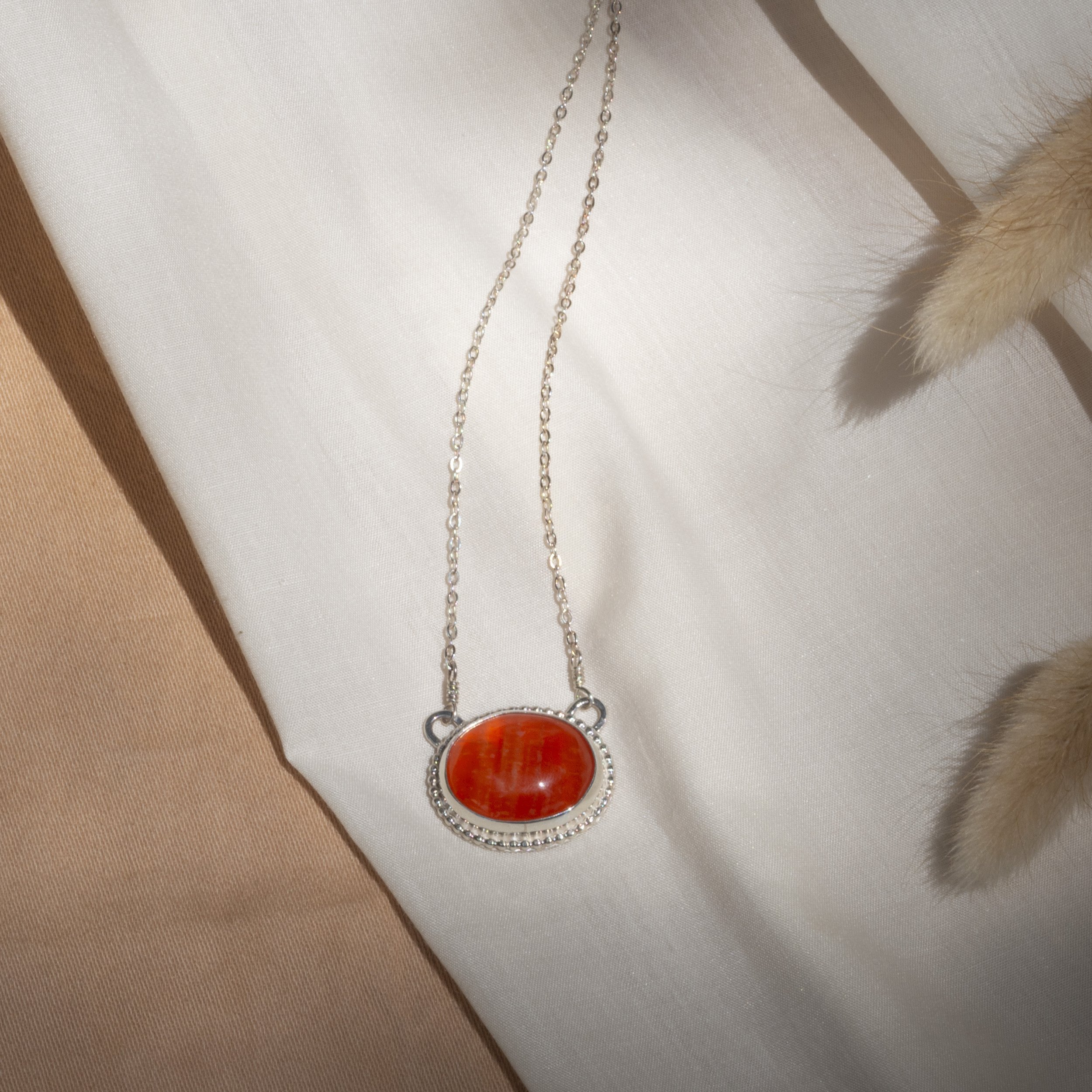 Silver necklace with an oval red stone pendant displayed on beige and light fabric background.