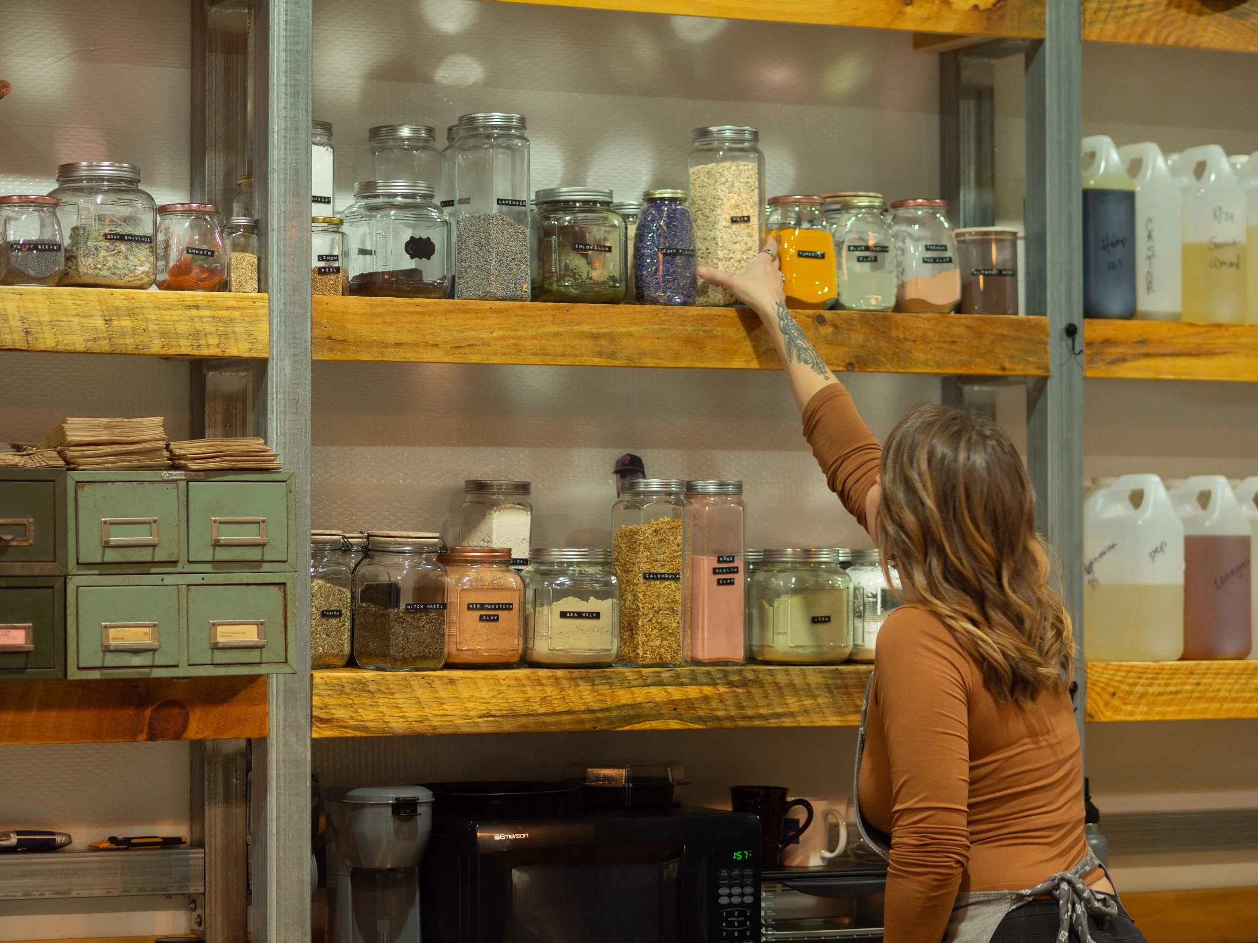A person reaching for a jar on a wooden shelf filled with various labeled jars containing different dry goods and ingredients. There are metal drawers on the left and plastic jugs on the right. The setting appears to be a kitchen or pantry.