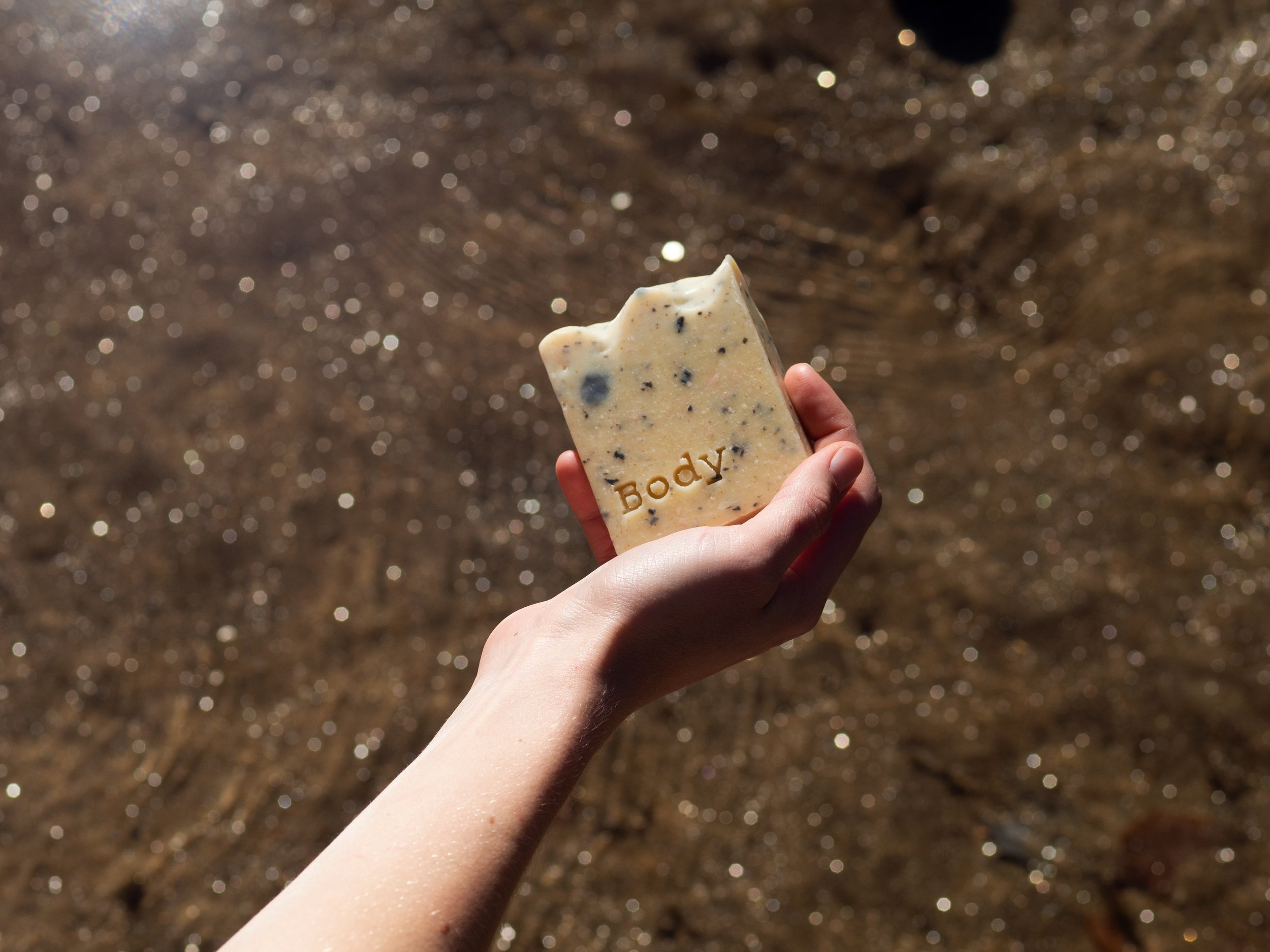 Hand holding a bar of soap with 'Body' engraved, outdoors with sunlight reflecting on water in the background.