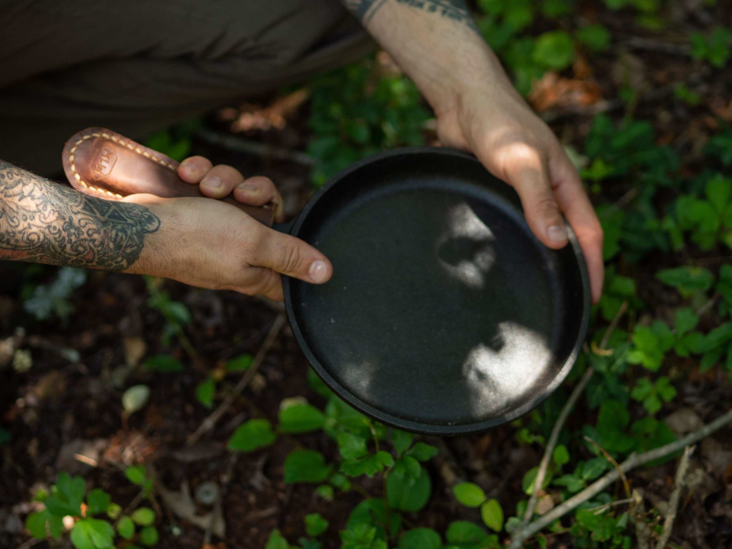 Person holding a black frying pan with leather handle in a forest setting, surrounded by green foliage.