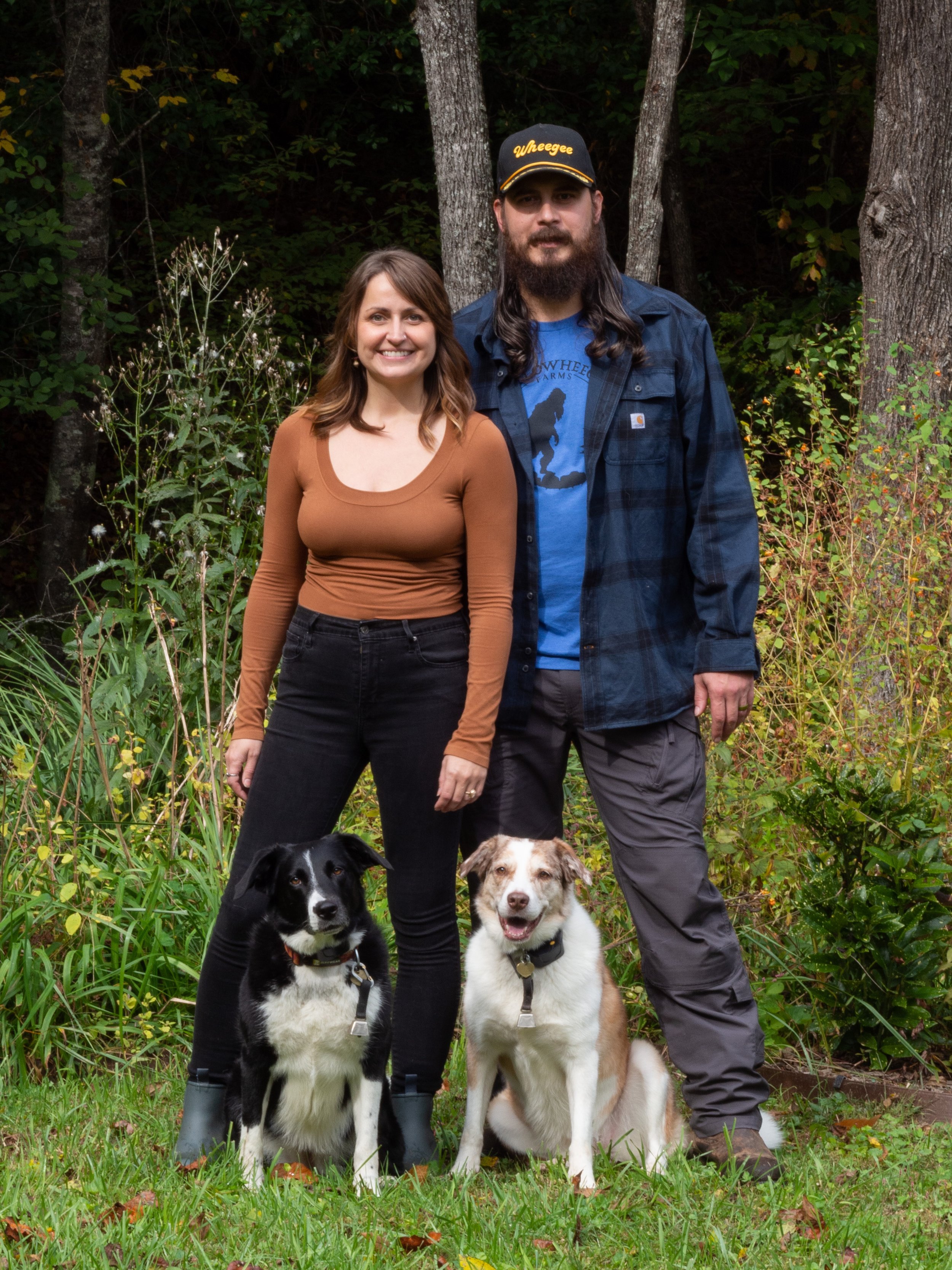 A man and woman standing together outdoors with two dogs sitting in front of them, surrounded by greenery.