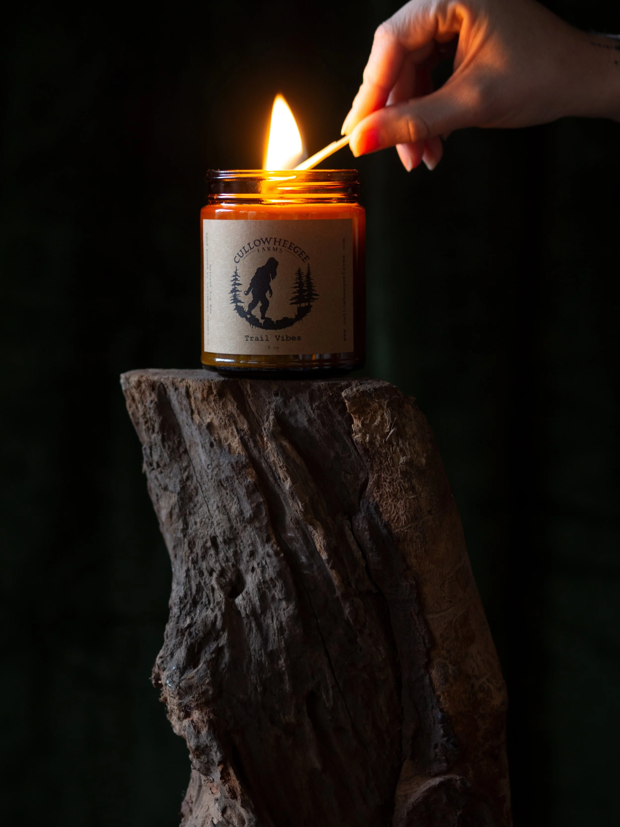A hand lighting an amber glass candle labeled "Cullowheegee Farms Trail Vibes" on a piece of wood.
