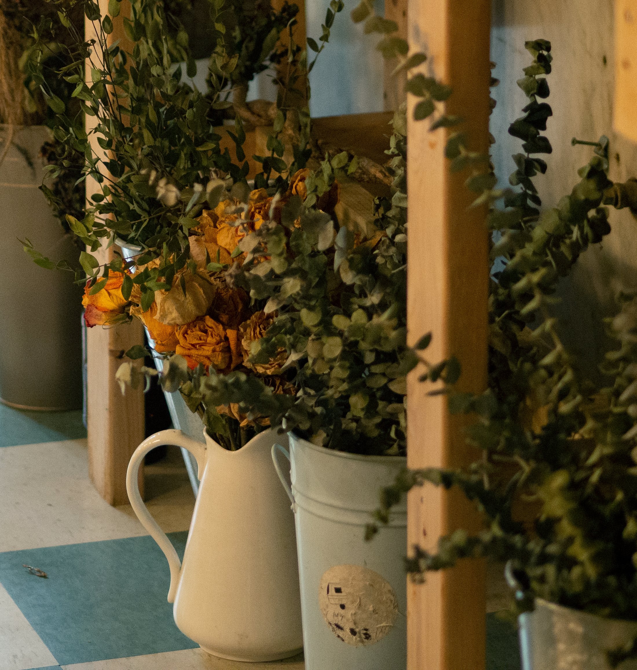 Flowers and greenery in a ceramic pitcher and metal bucket on a shelf.