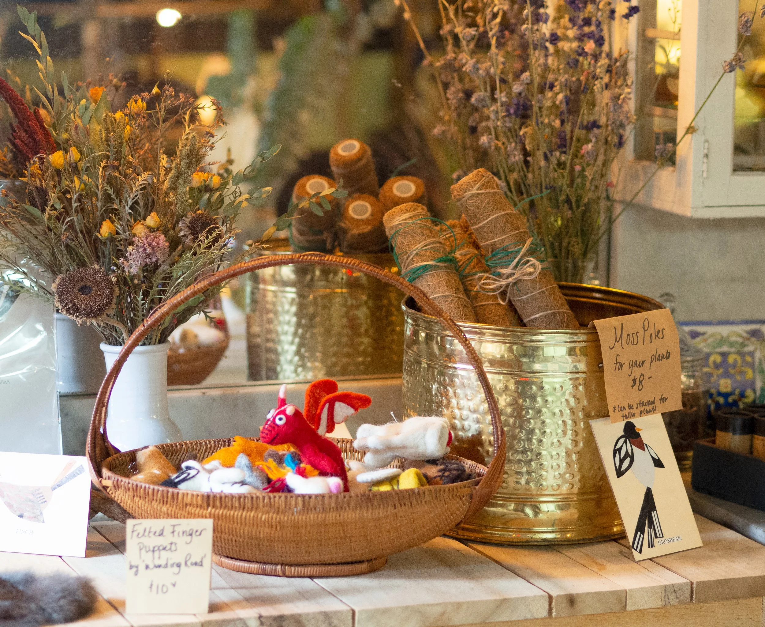 Display of dried flowers, woven basket with felted finger puppets, and metal container with moss poles for sale in a store.