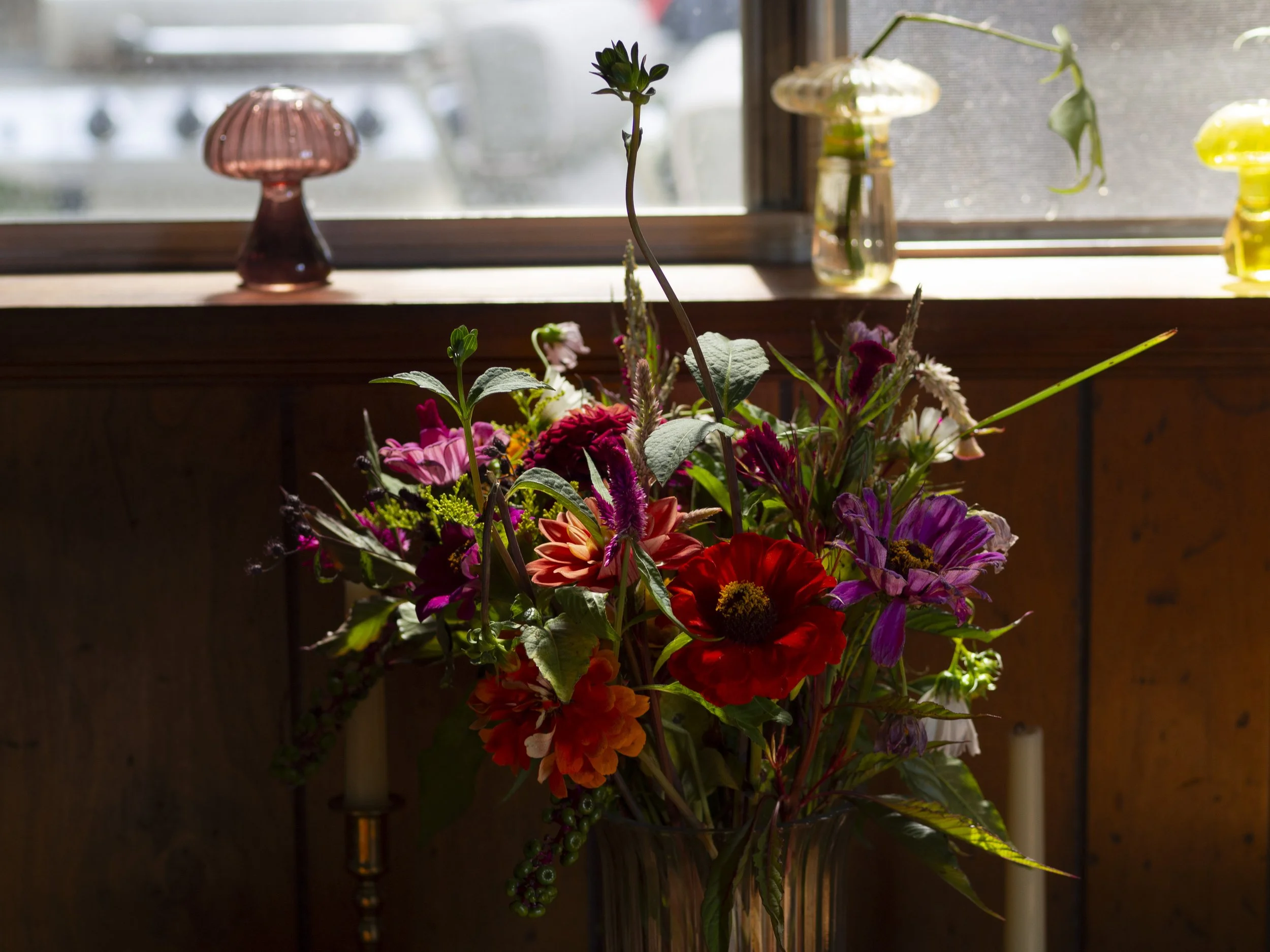 A colorful bouquet of flowers, including red, purple, and orange blooms, placed in a glass vase indoors near a window. Two glass mushroom-shaped decorations are visible on the windowsill.