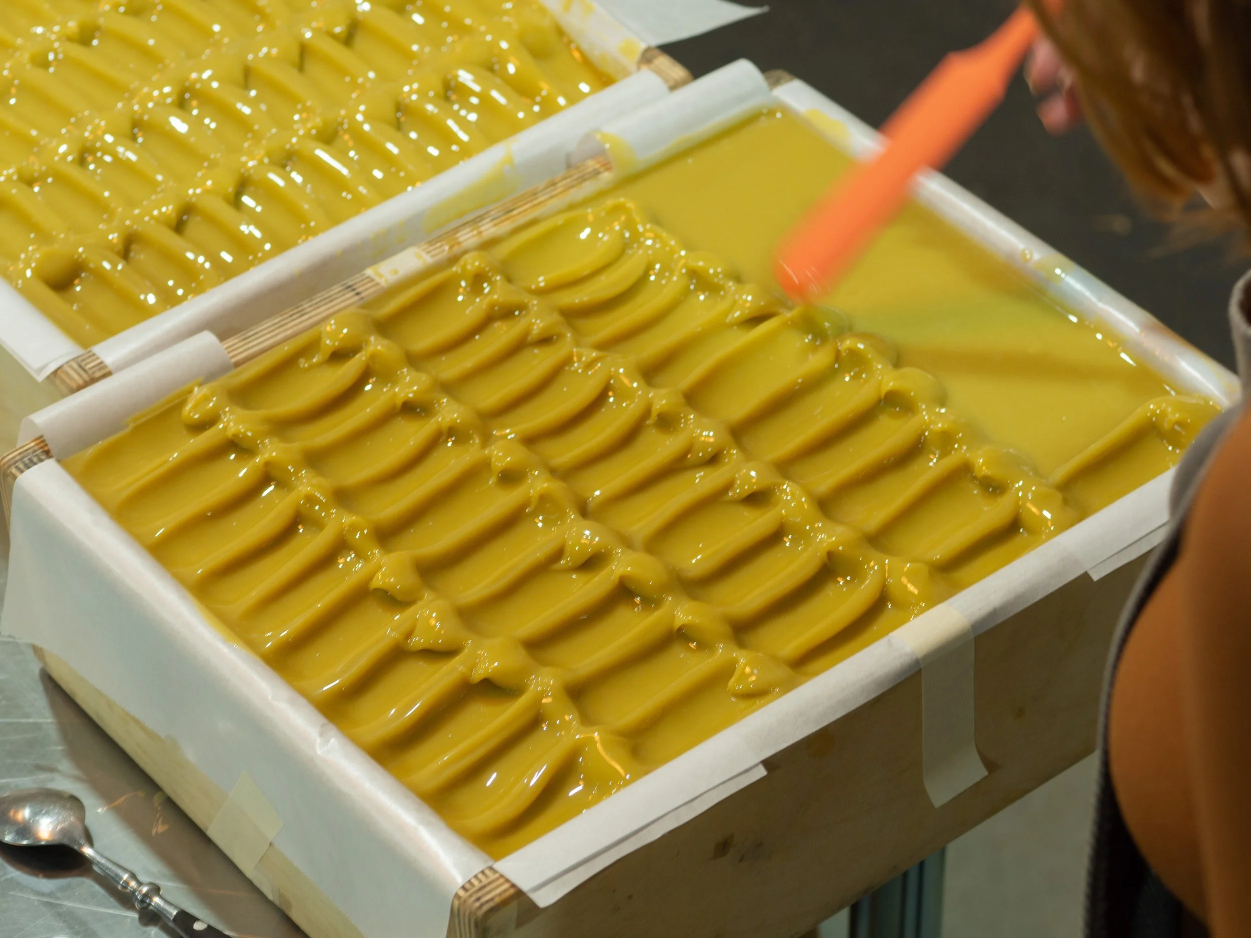 Person using a spatula to spread yellow soap batter in a wooden mold lined with parchment paper.