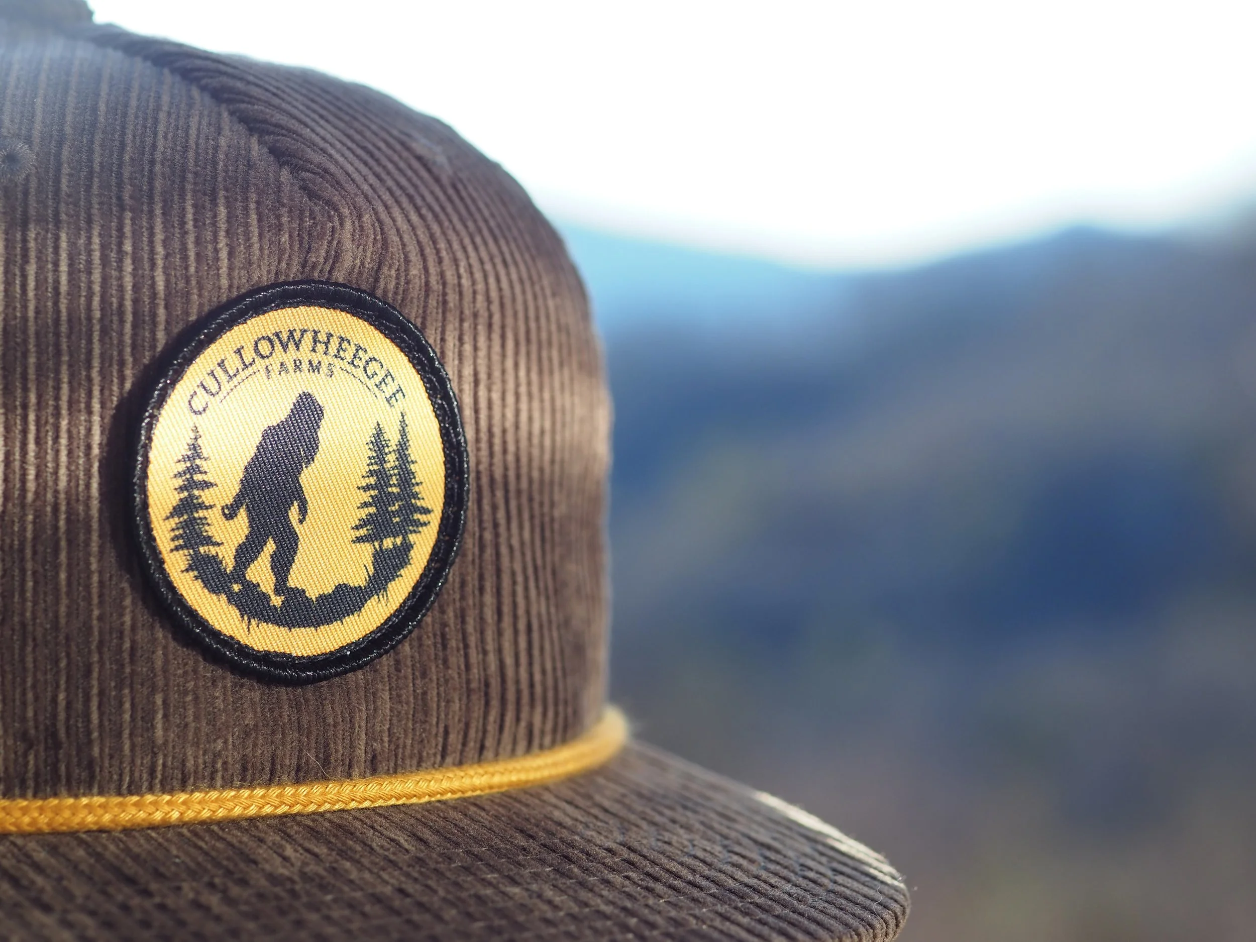 Close-up of a brown corduroy cap with a circular patch featuring a silhouette of Bigfoot and trees, labeled "Cullowheegee Farms."