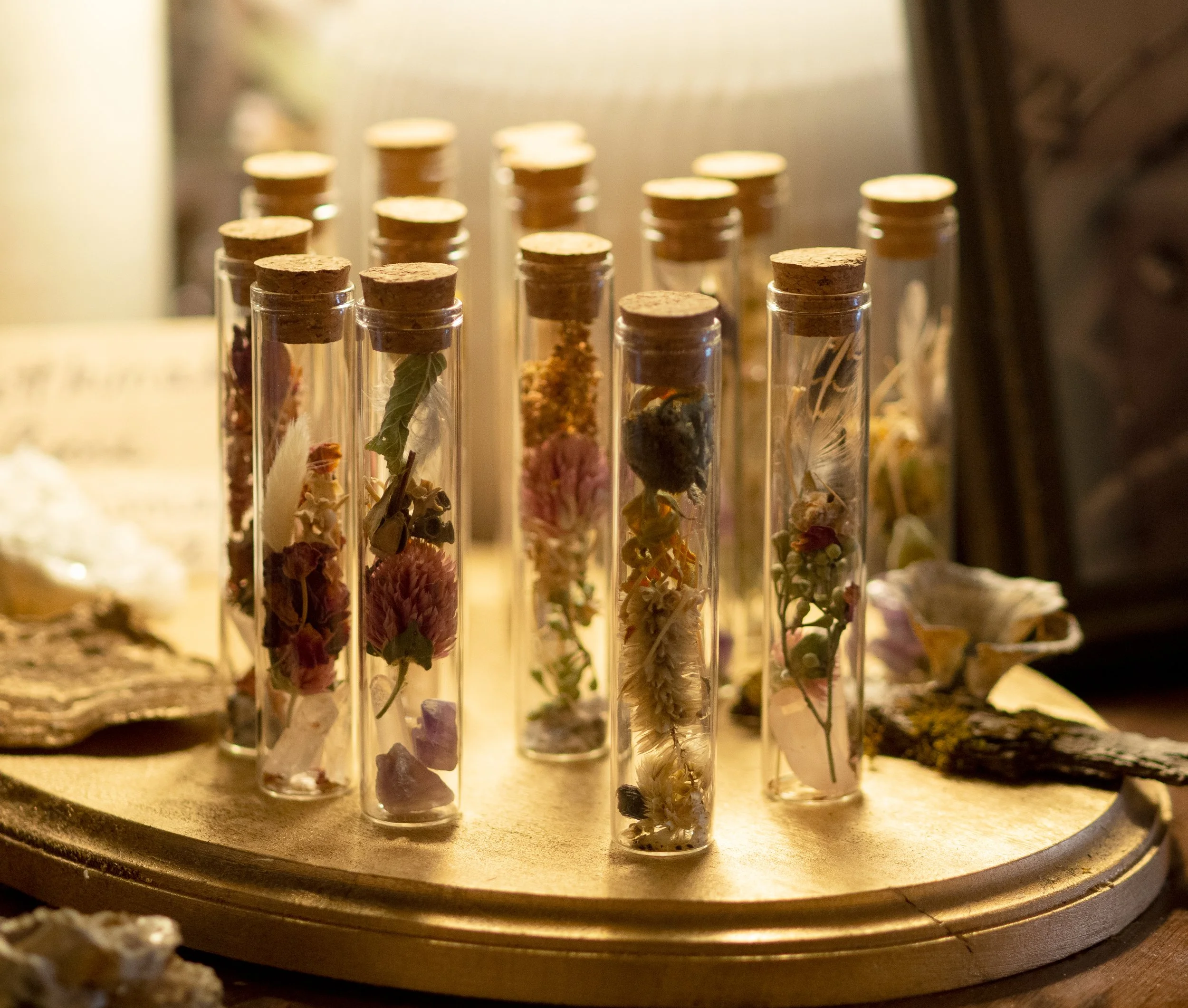 Glass vials with cork stoppers containing dried flowers and herbs, arranged on a round gold tray.
