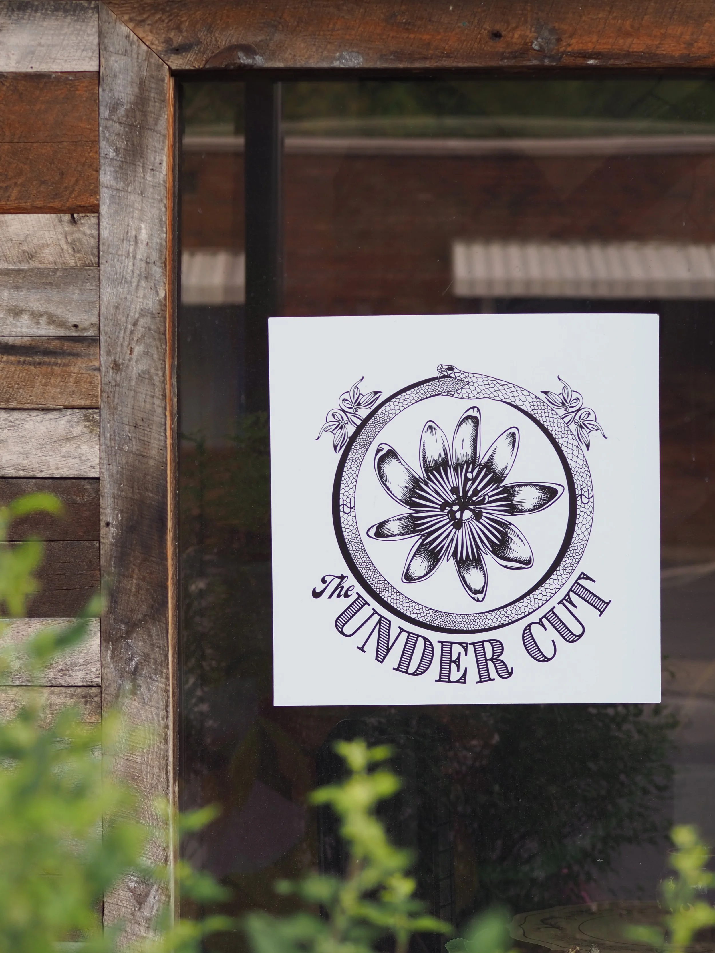 Sign or poster on a wooden-framed window displaying the logo and name "The Under Cut" with a circular design featuring a flower and a snake.