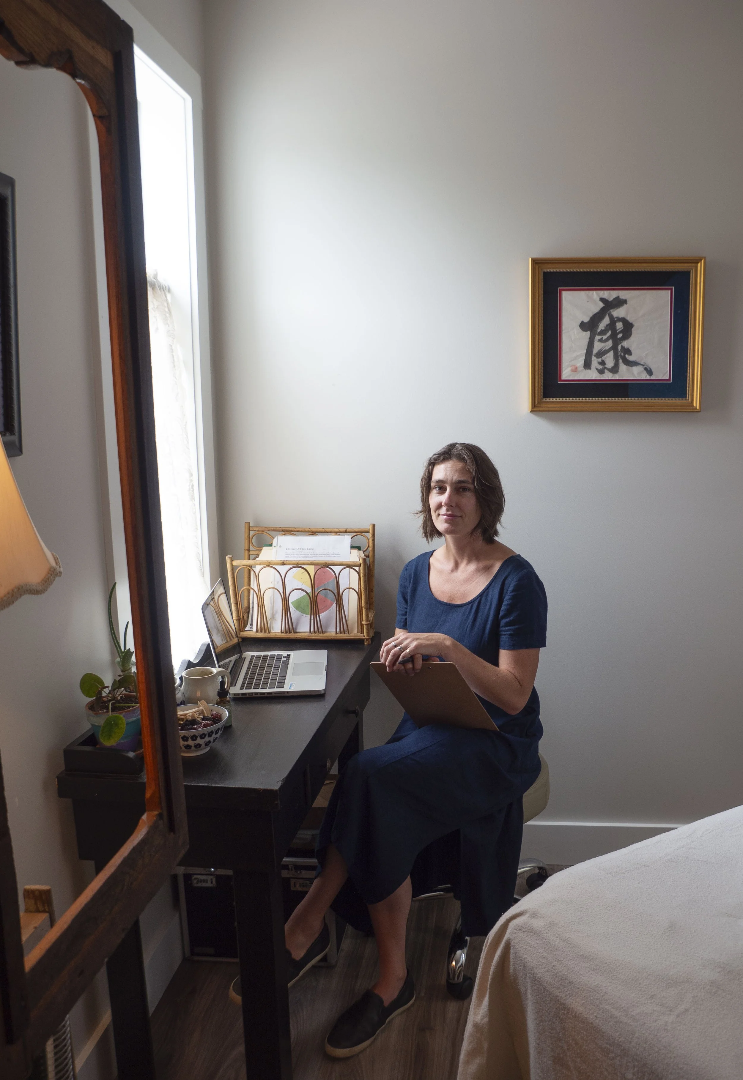 A person sitting at a desk holding a clipboard, with a laptop, plants, and framed artwork on the wall in a cozy, well-lit room.