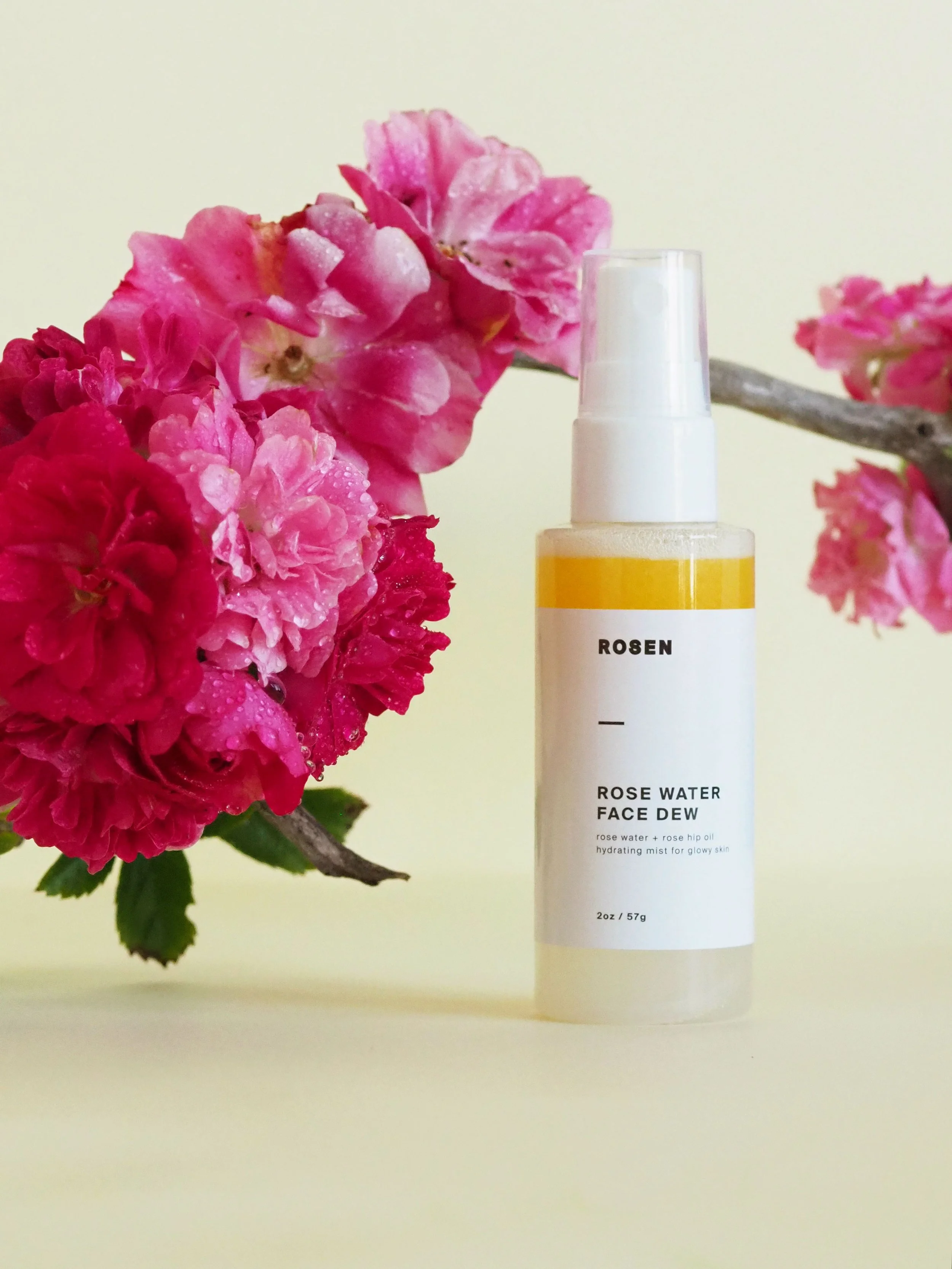 A bottle of Rosen Rose Water Face Dew next to bright pink flowers on a light background.