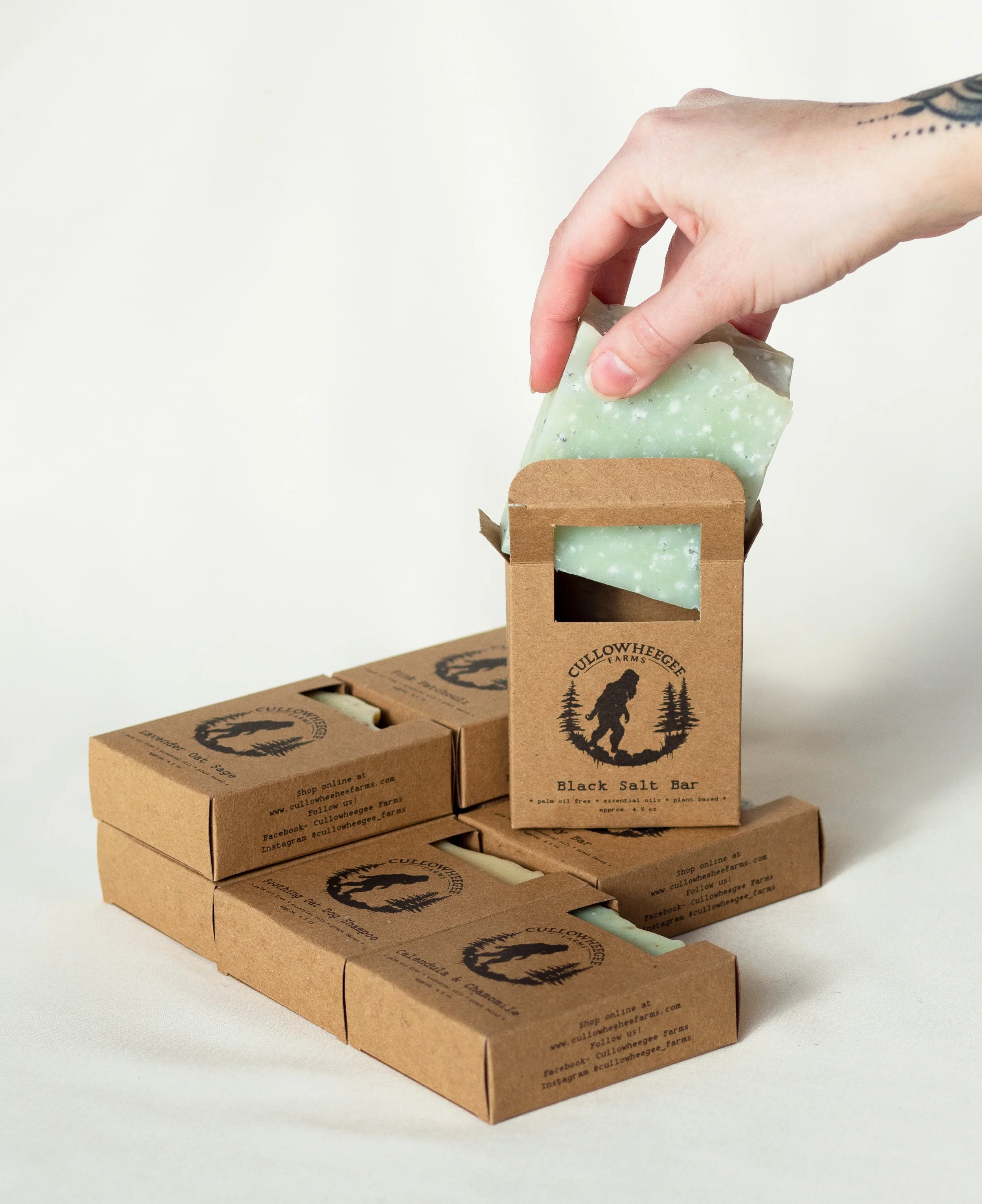 Soap bars in brown cardboard packaging with hand removing one bar, featuring wilderness-themed design and branding.