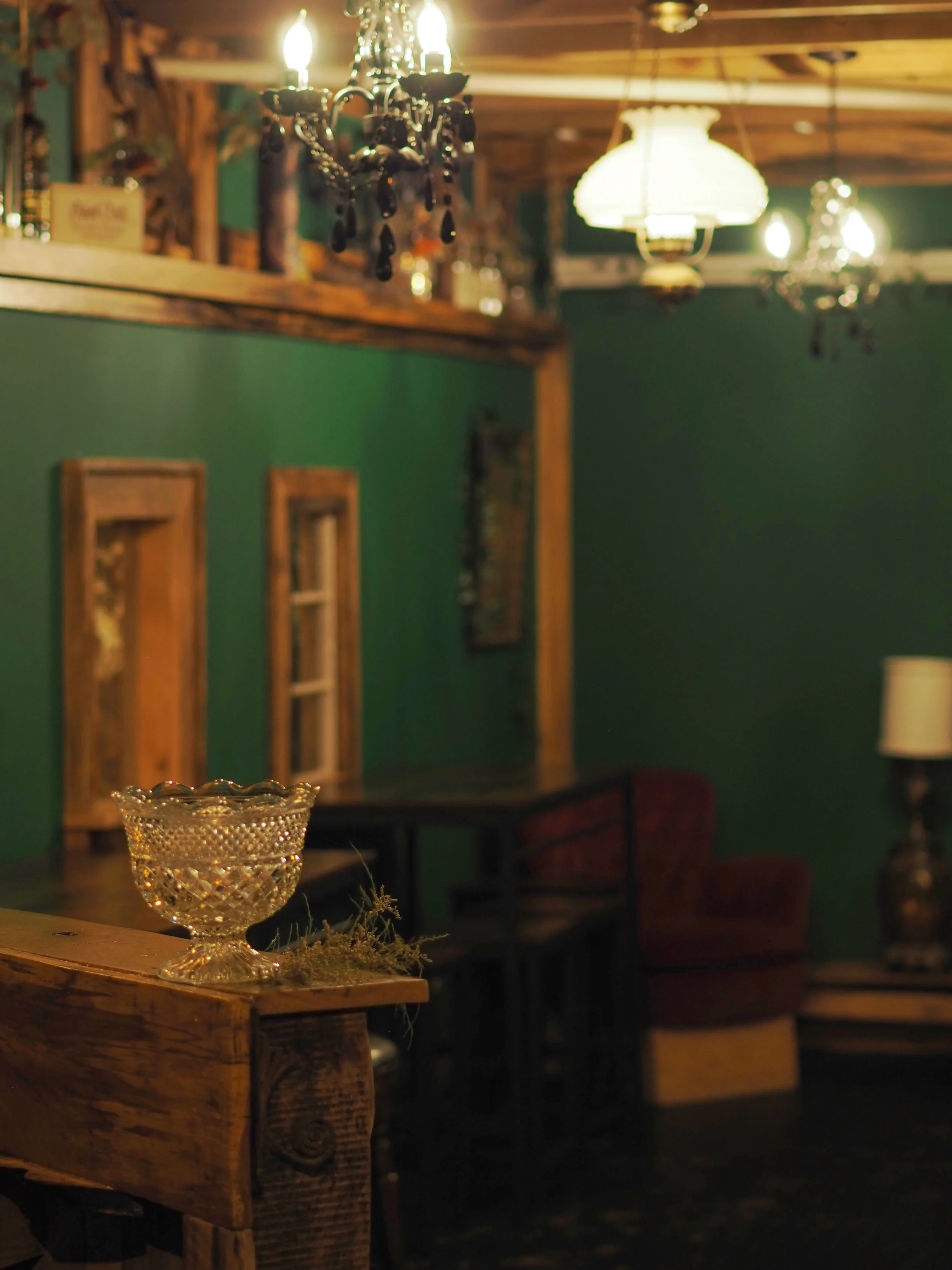 Vintage-style room with dark green walls, wooden accents, a crystal bowl on a wooden table, chandeliers with black beads, and a red velvet chair in the background.