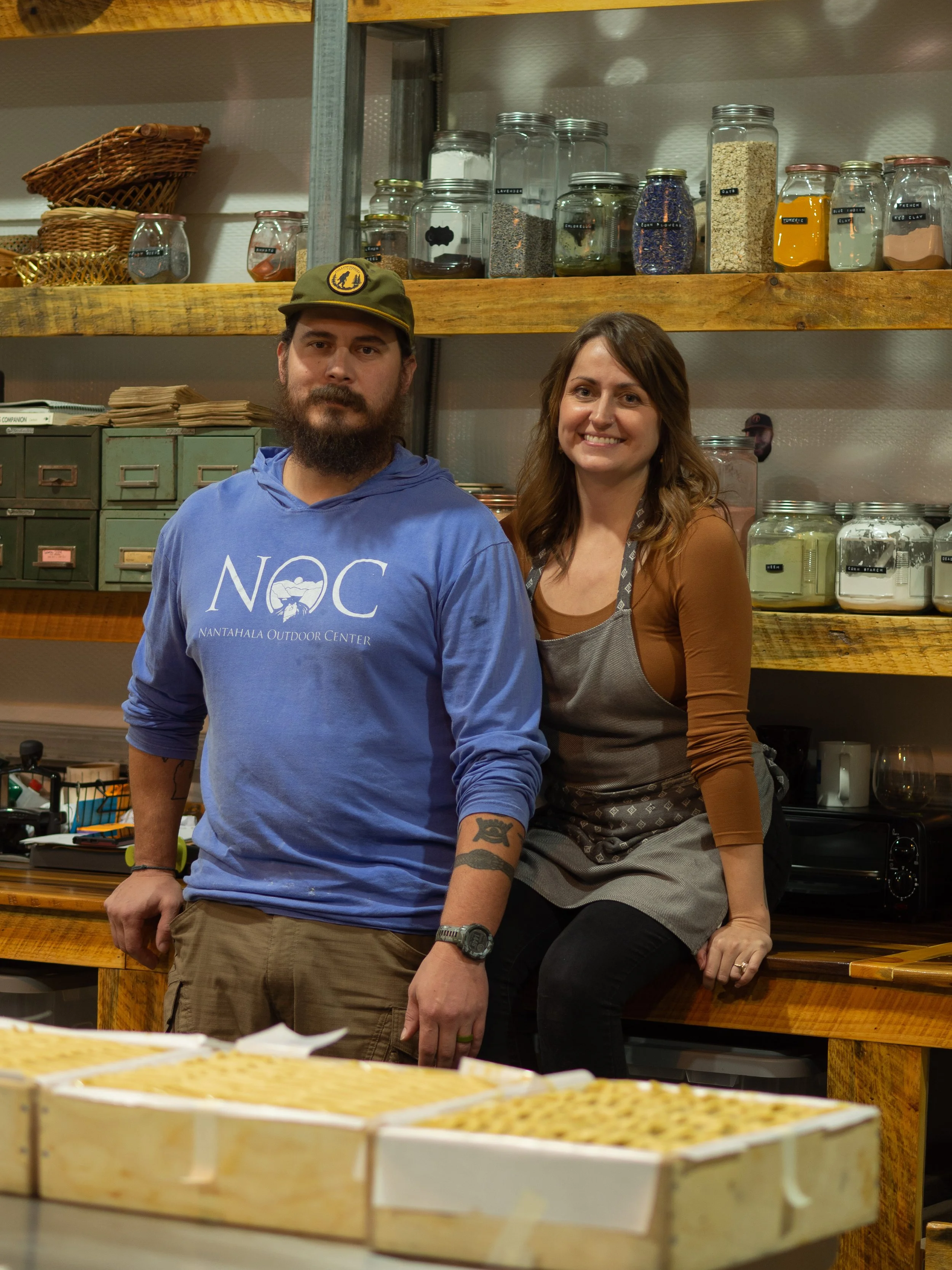Two people standing in a kitchen with shelves of jars and baskets, and trays of food in the foreground.
