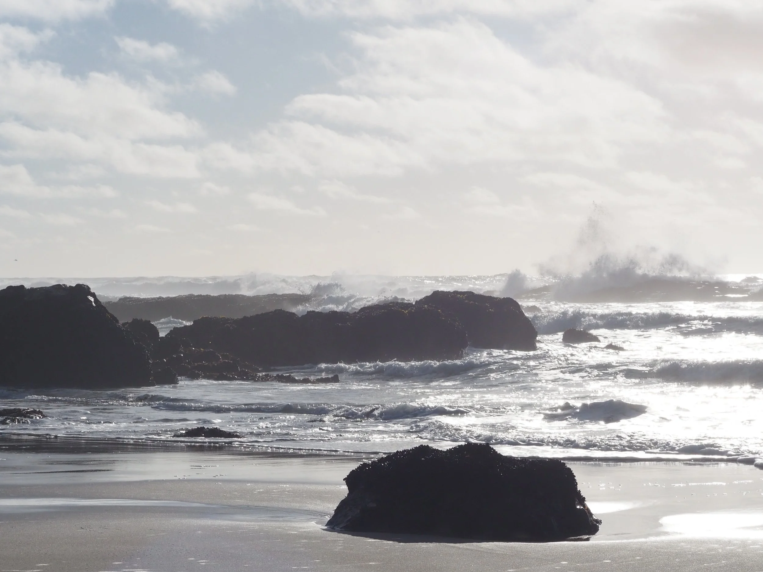 Waves crashing against large rocks on a beach, with wet sand and cloudy sky.